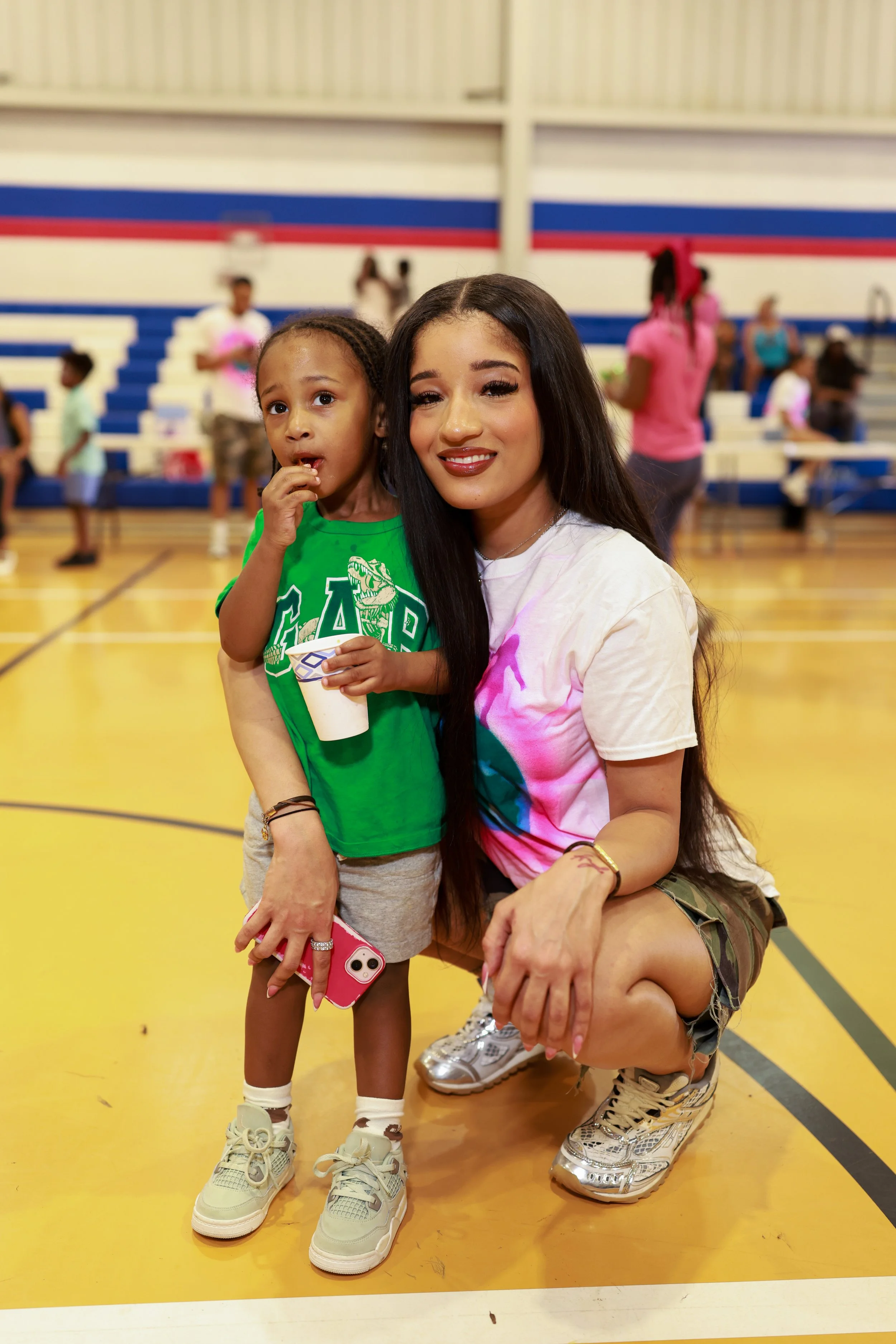 A woman and a young girl posing together in a gymnasium. The girl is wearing a green shirt, gray shorts, and sneakers, and holding a cup of ice cream and a pink phone. The woman is squatting, wearing a colorful T-shirt, camouflage shorts, and metalli