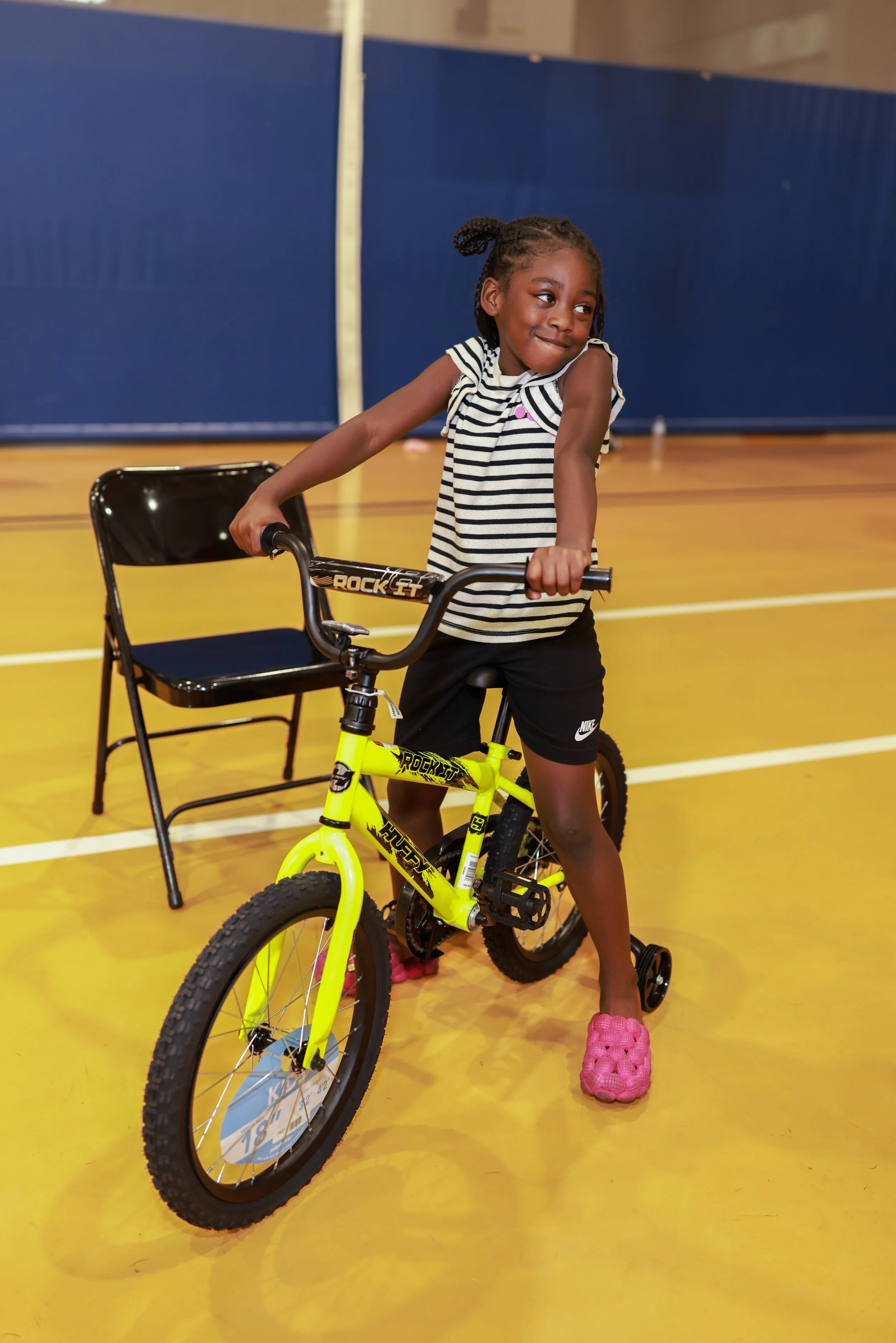 A young girl stands in a gymnasium holding a yellow bicycle, wearing a striped shirt, black shorts, pink Crocs, and has braided hair. There is a black folding chair next to her and a blue divider in the background.