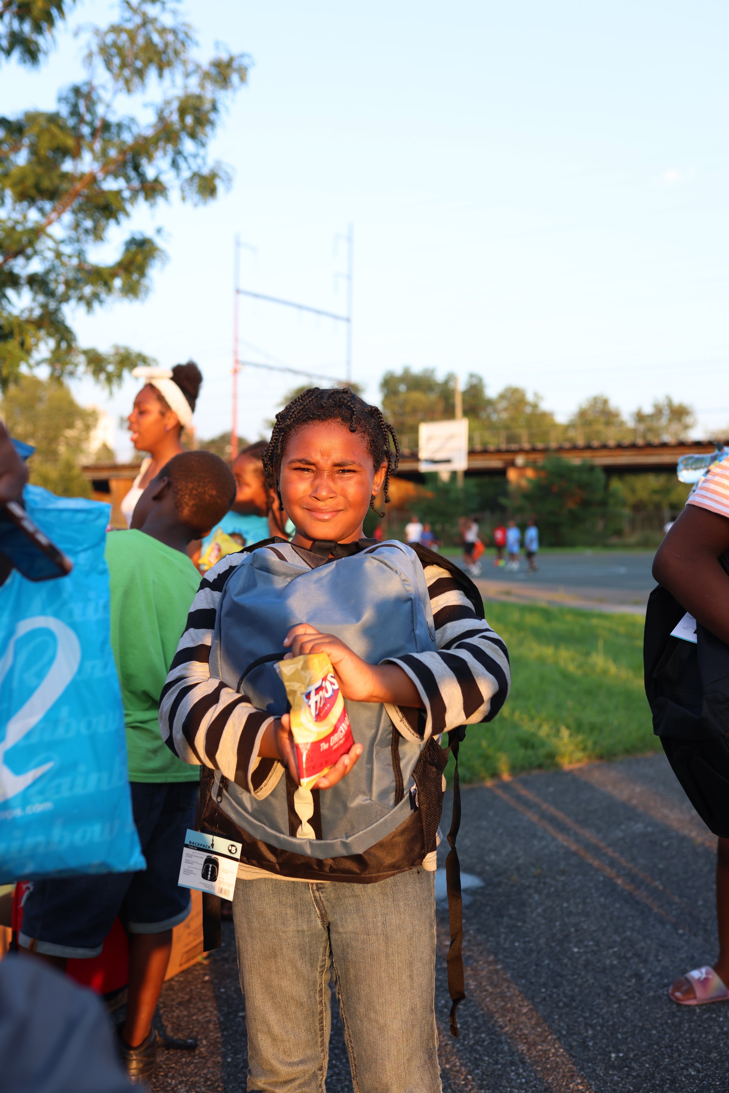 Young girl with braided hair and striped shirt holding snacks, standing outdoors during the daytime with other children in the background.