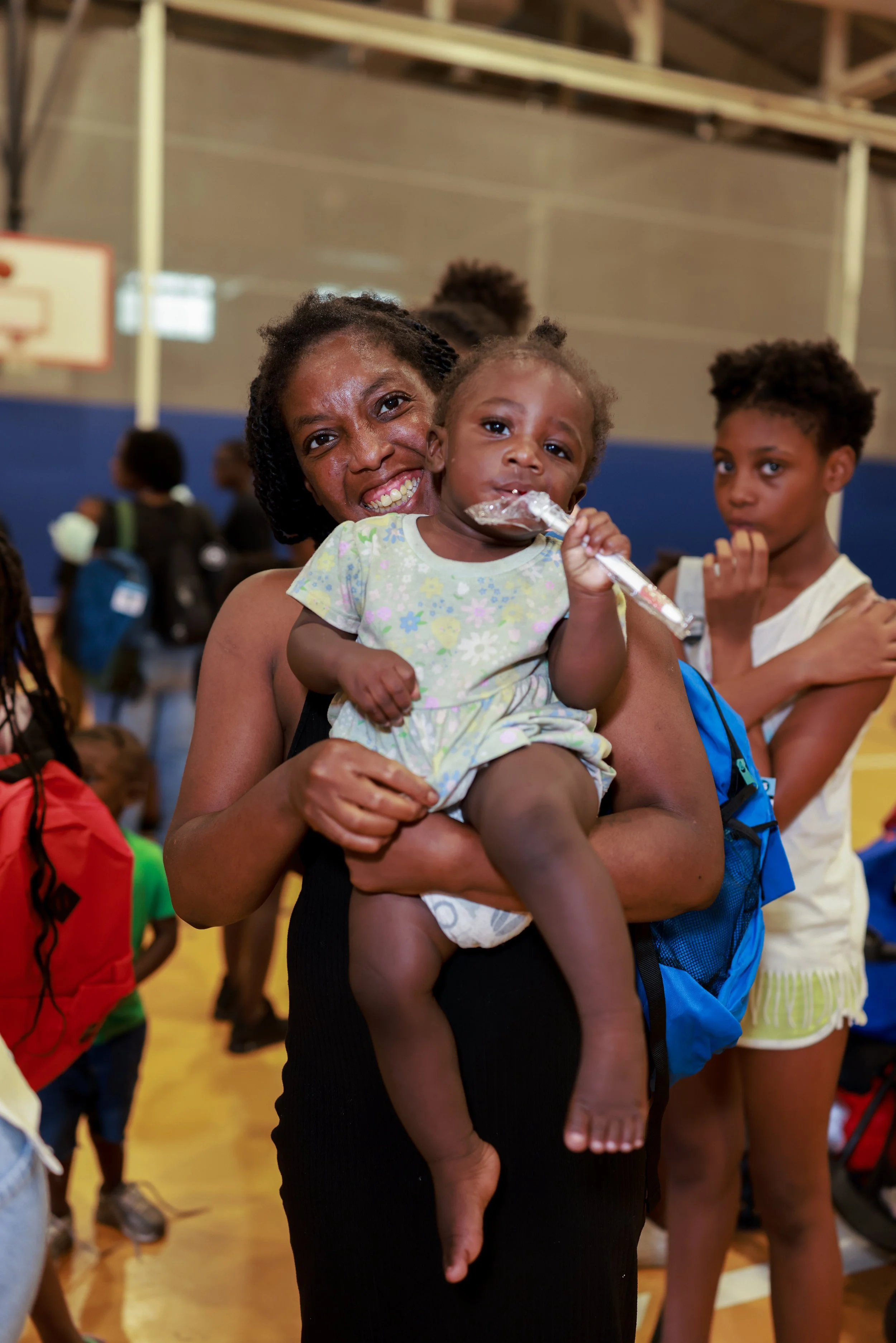 A woman holding a young child in a gymnasium with children in the background.