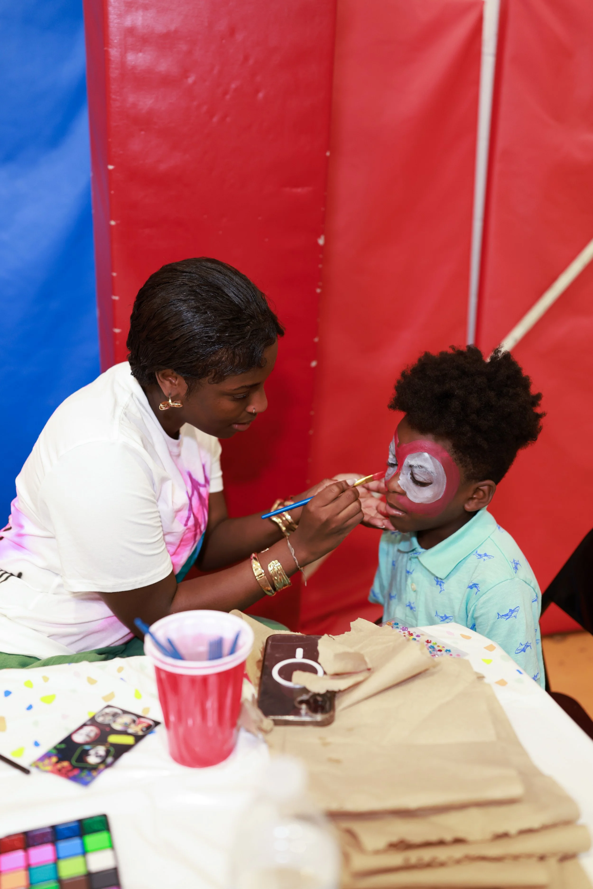 A woman is painting a face white with pink and red accents on a child's face during a celebration or event, with various craft supplies and decorations on a table.