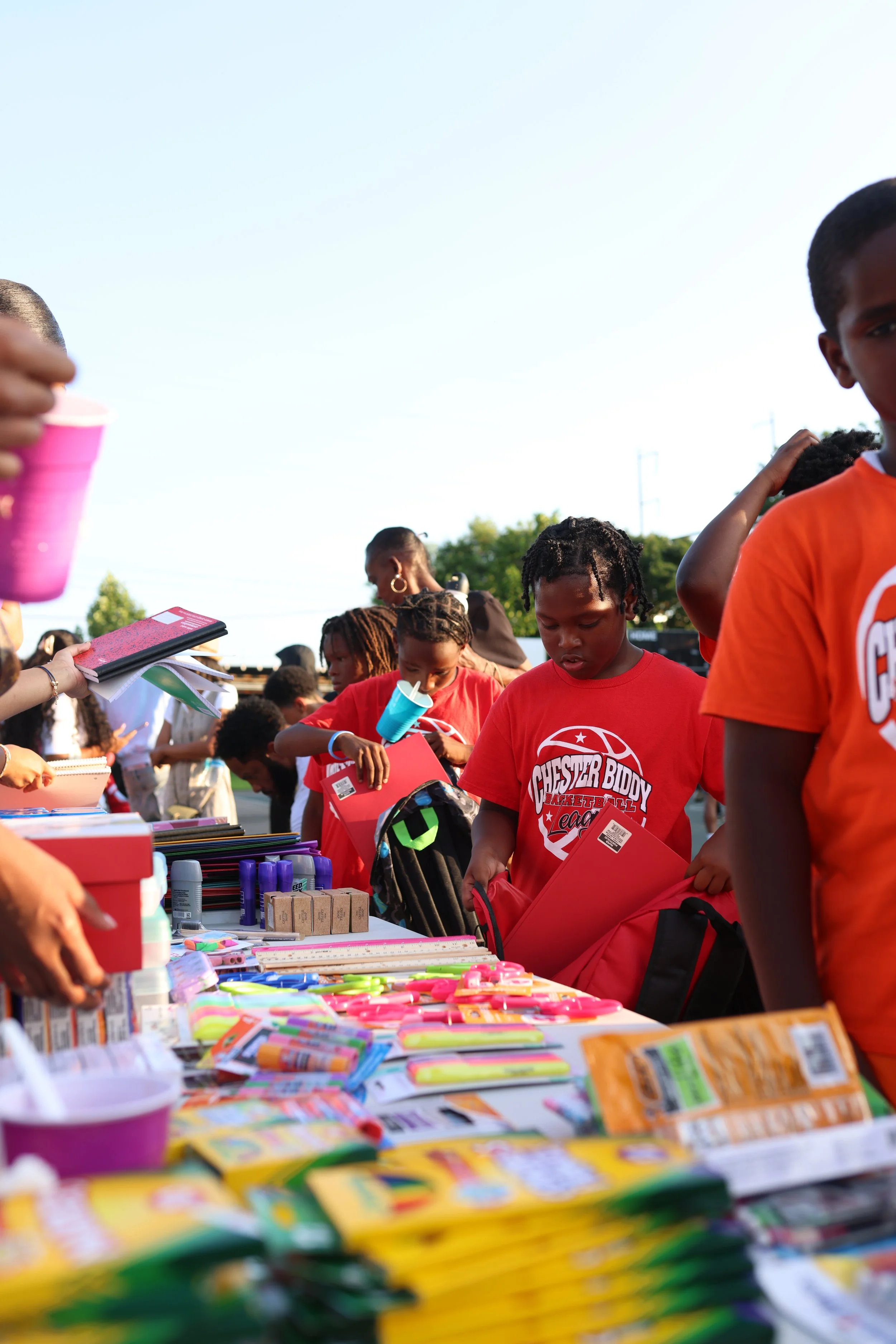 Group of children and adults at an outdoor school supply giveaway, with tables full of colorful school supplies like notebooks, pens, and markers.