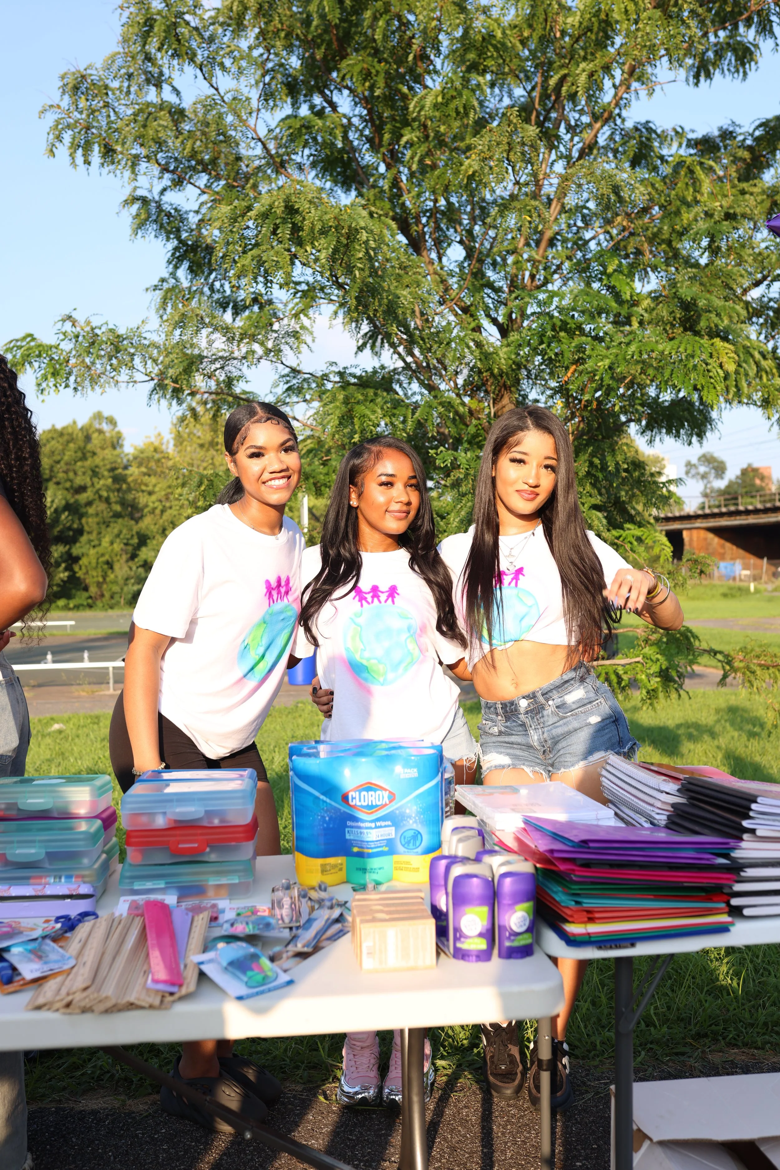 Four young women wearing white t-shirts with the earth and pink figures printed on them are standing behind a table outdoors. The table is filled with various school supplies, including notebooks, folders, pens, and disinfecting wipes. They are smili