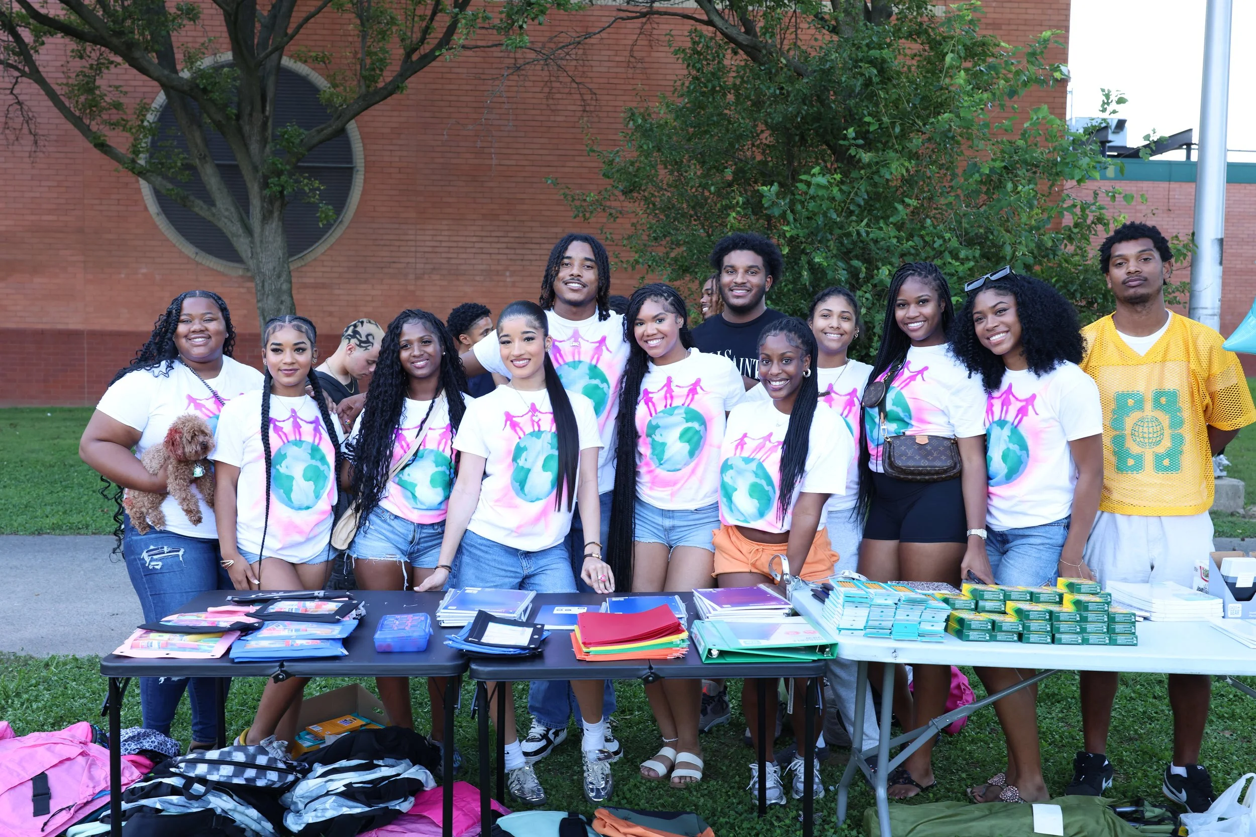 A group of diverse young people smiling and standing behind a table with school supplies at an outdoor event, with a brick building, trees, and a pole in the background.