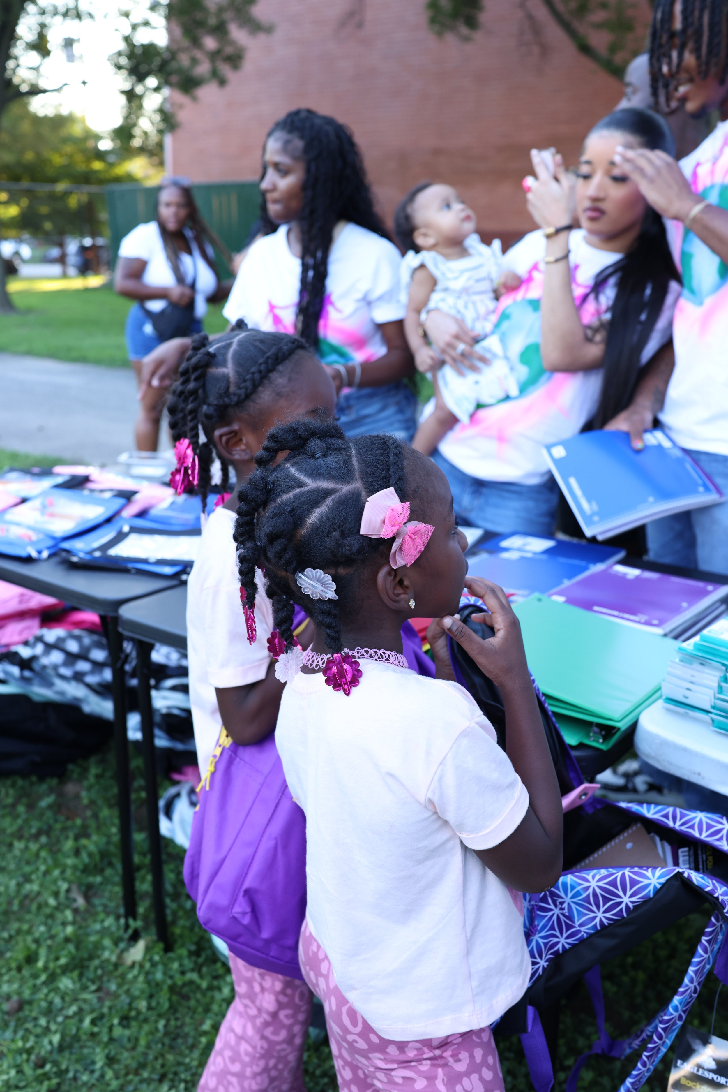 Several children and young women gathered around tables with books, notebooks, and backpacks outdoors on a grassy area, participating in an event or educational activity.