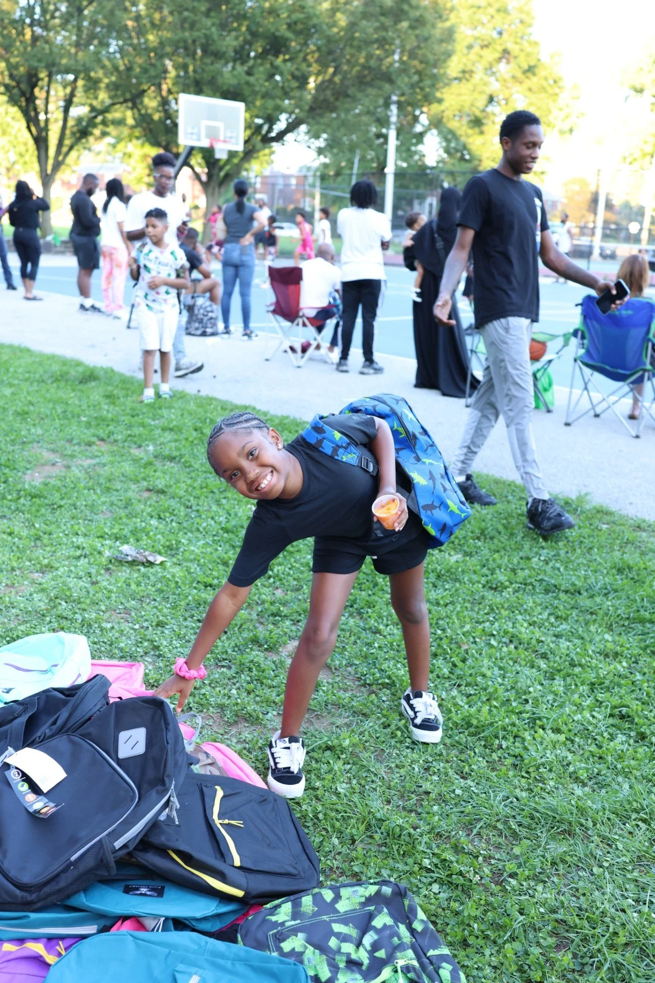A young girl with a backpack and a pink scrunchie, smiling and crouching on the grass, surrounded by backpacks and bags, at an outdoor gathering with people, trees, and a basketball court in the background.