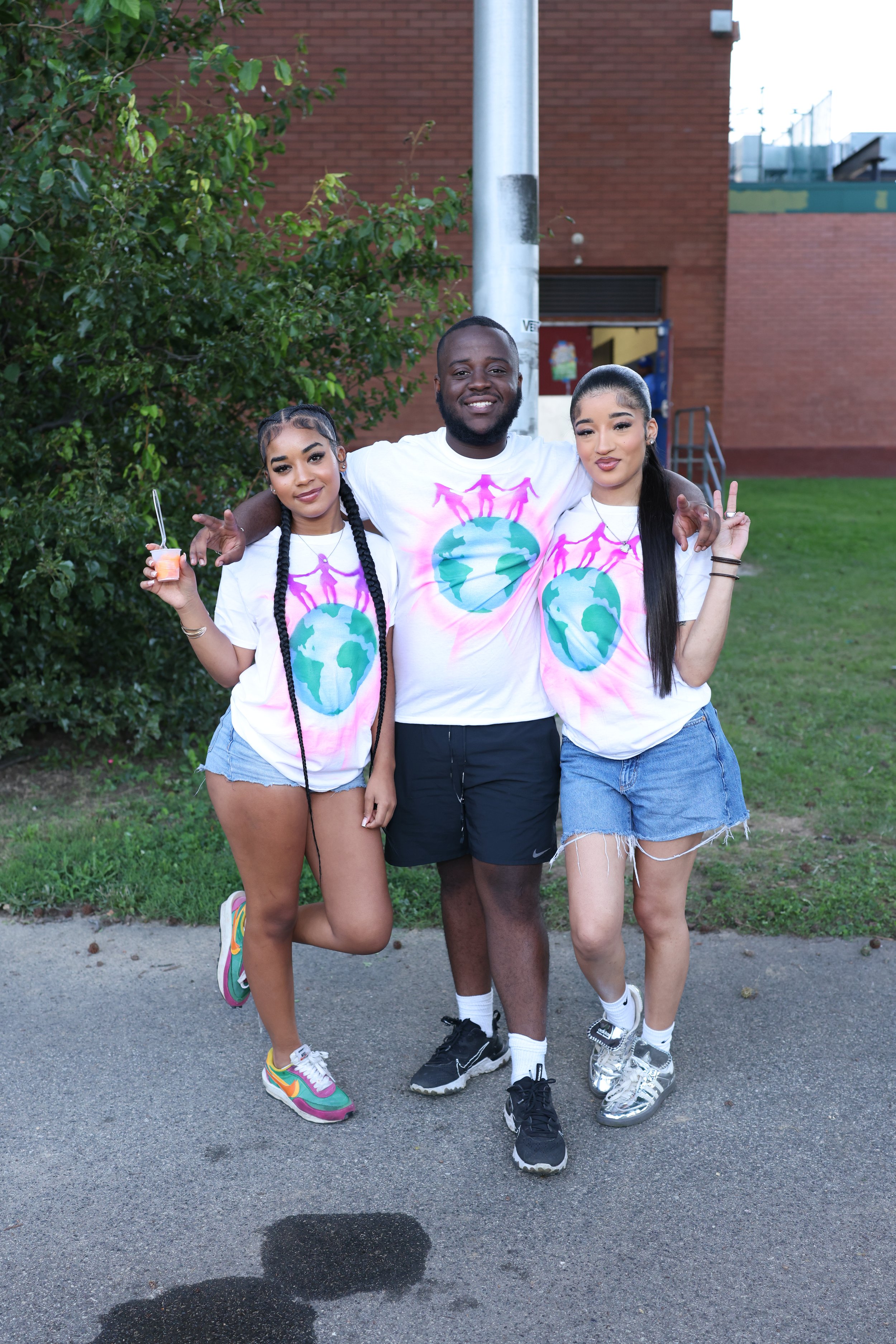 Three young adults standing on a sidewalk in front of a brick building and greenery, wearing matching white T-shirts with a globe and people holding hands design, smiling and posing for the photo.