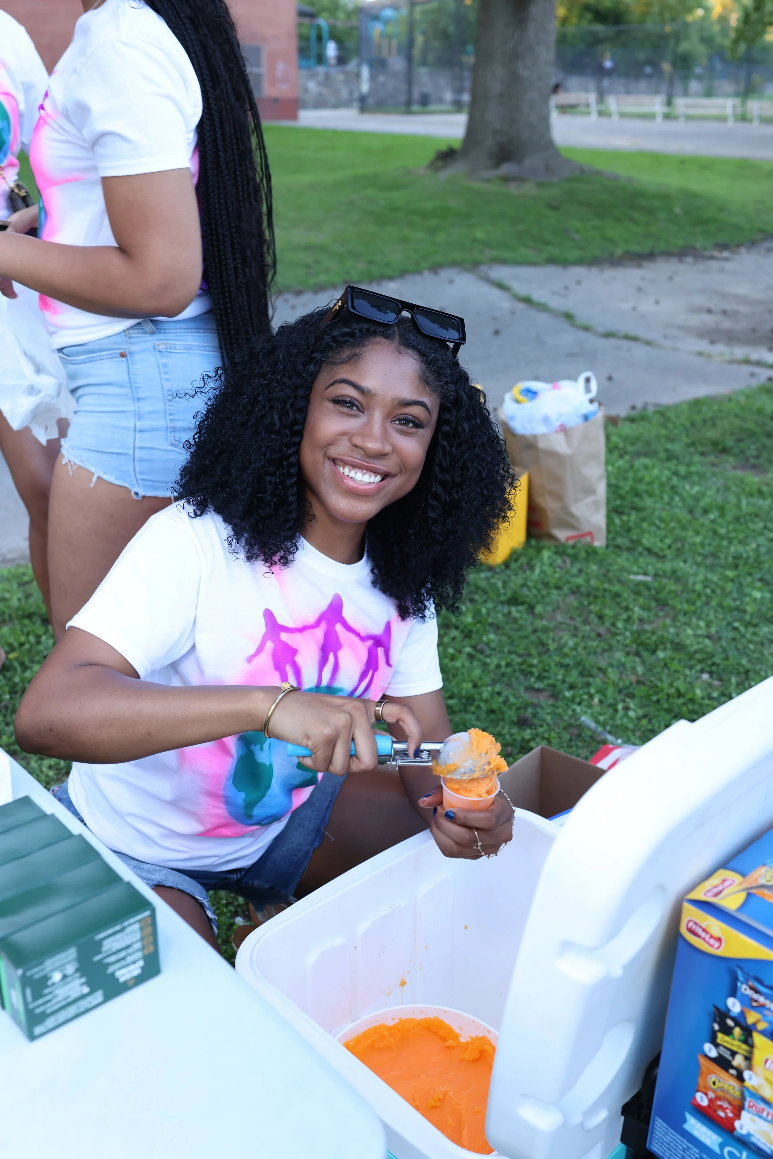 Young woman with curly black hair and sunglasses on her head, smiling while scooping orange ice cream into a cone at an outdoor event, with other people and green park background.