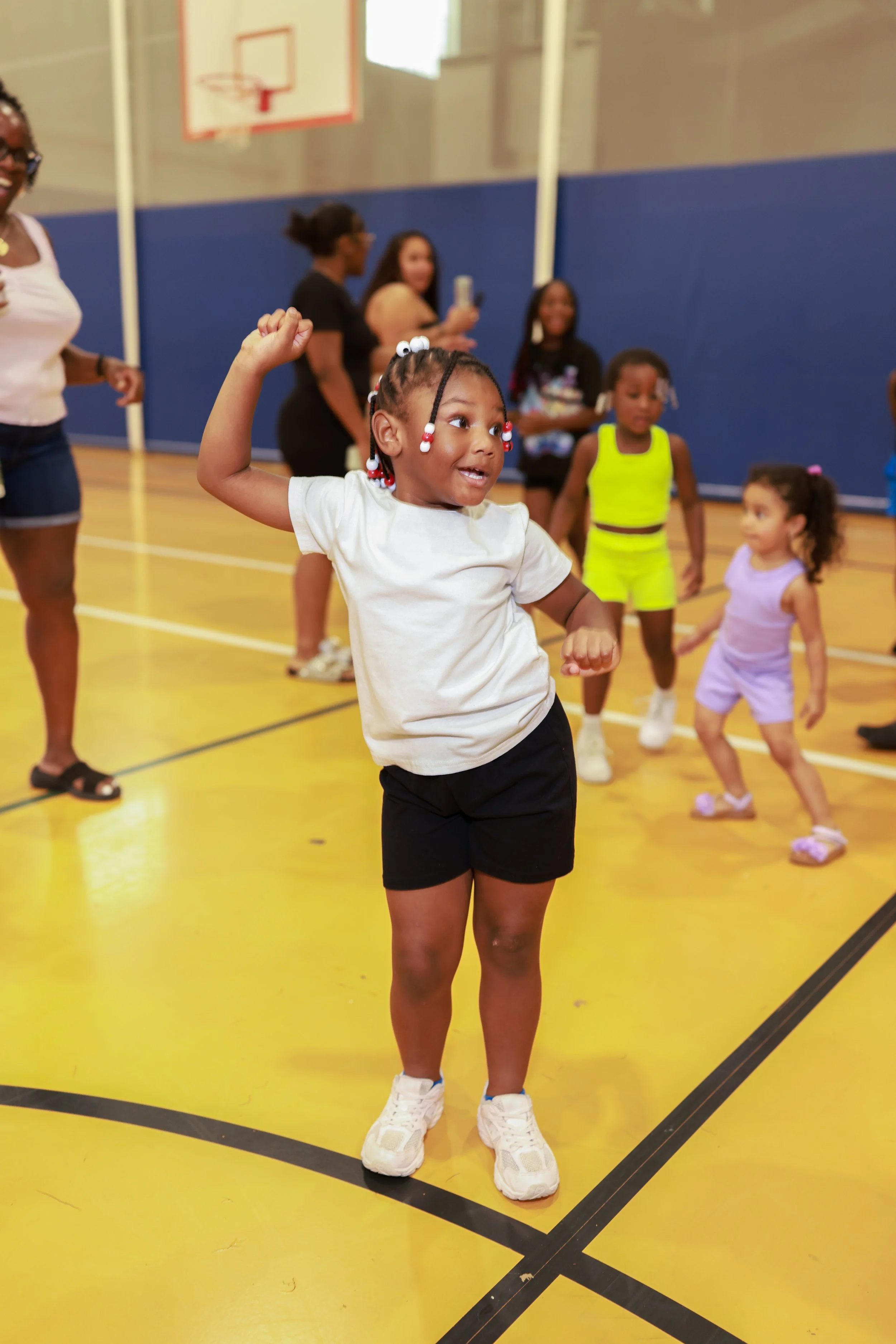 A young girl with braided hair and decorative beads is dancing in a gymnasium. Several children and adults are in the background, some watching and others talking. The gym has blue and yellow walls and a basketball hoop.