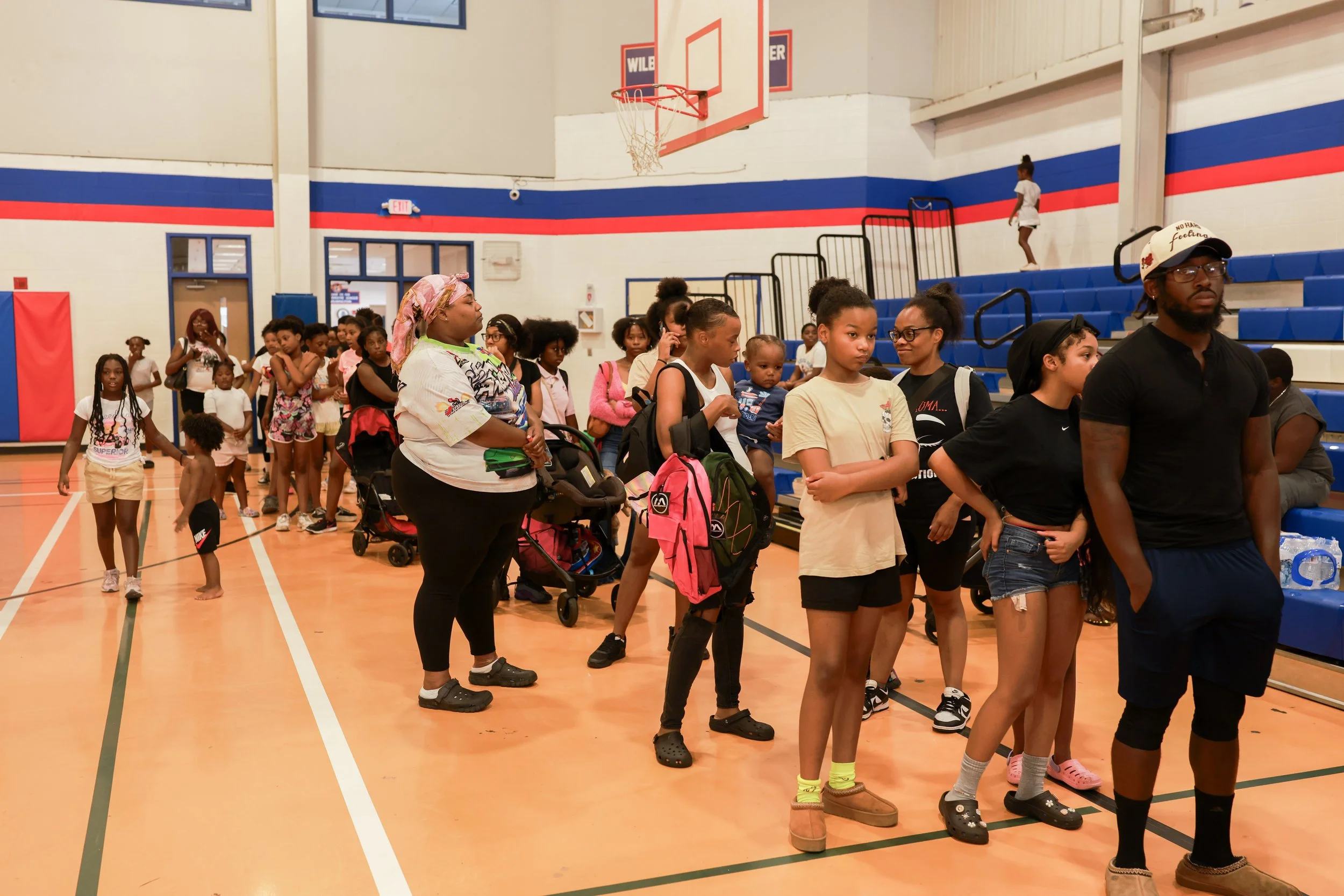 Children and adults standing in line inside a gymnasium with blue, white, and red painted walls and a basketball hoop in the background