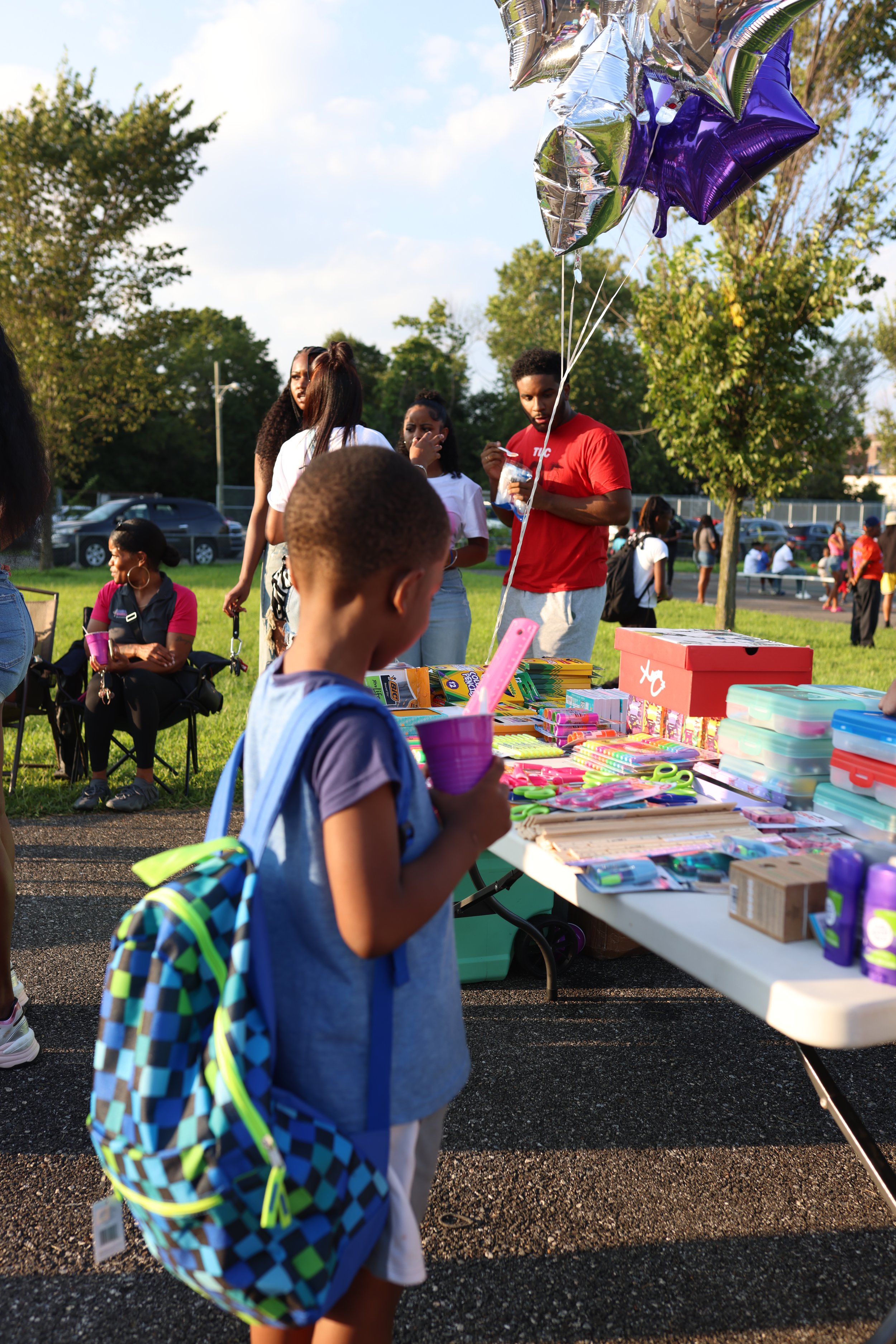 A young boy with a blue backpack looking at a table filled with school supplies, at an outdoor event. There are balloons in the air, and people in the background, some sitting and some standing, in a park with trees and cars.