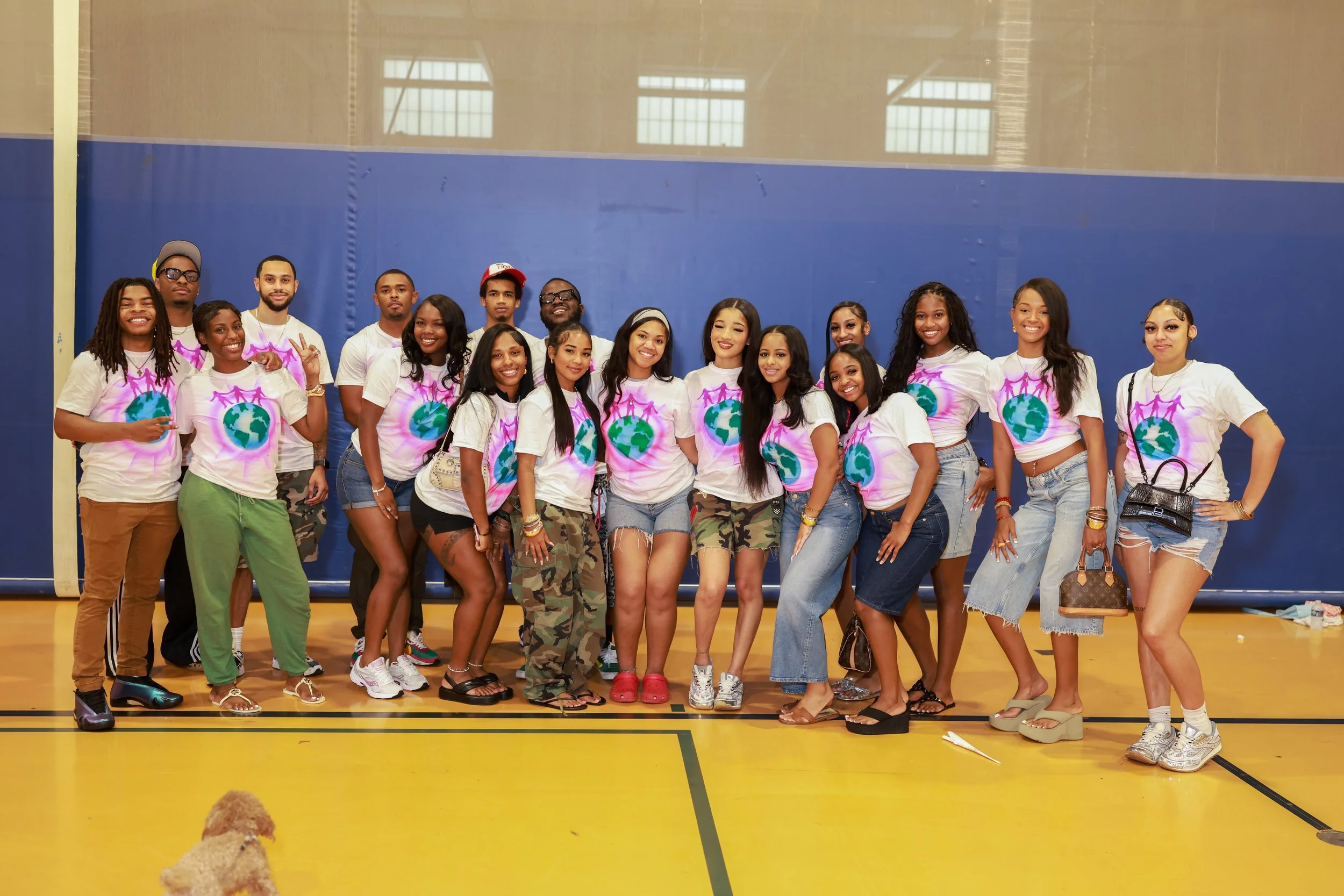 A diverse group of people standing together in a gymnasium, wearing matching t-shirts with a globe and people holding hands design, smiling at the camera.
