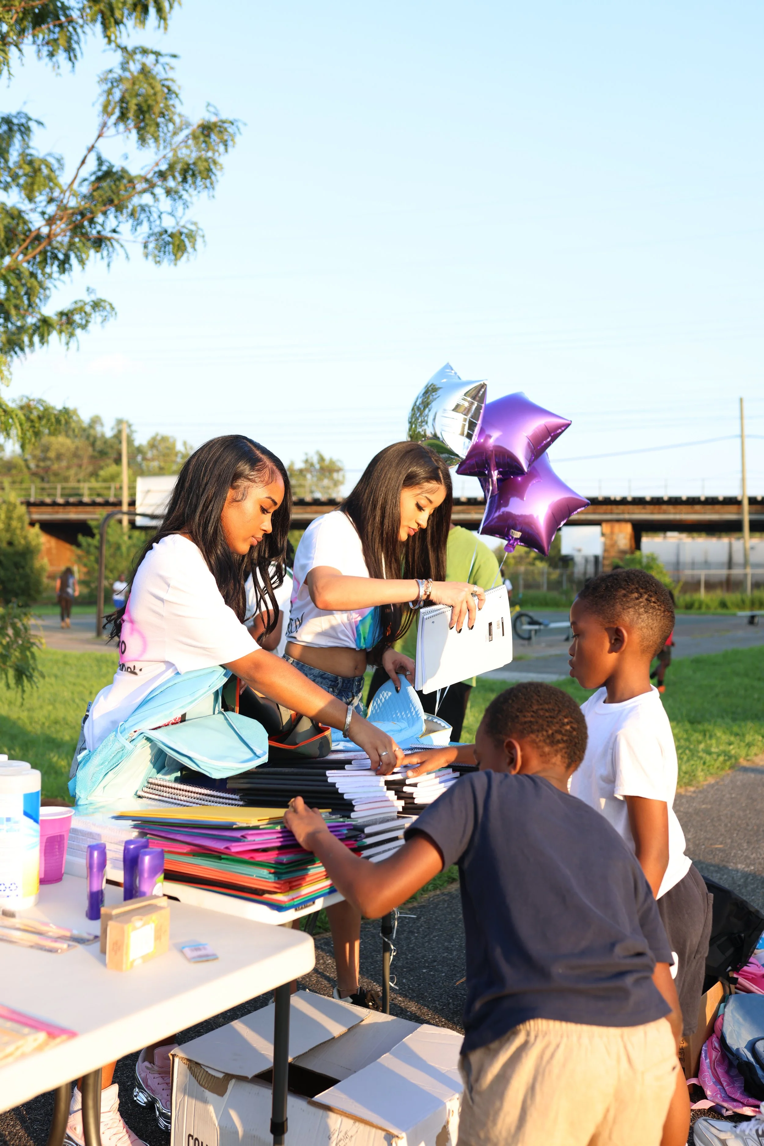 A group of children and teenagers standing around a table with school supplies and notebooks, outdoors during daytime with balloons and a bright sky.