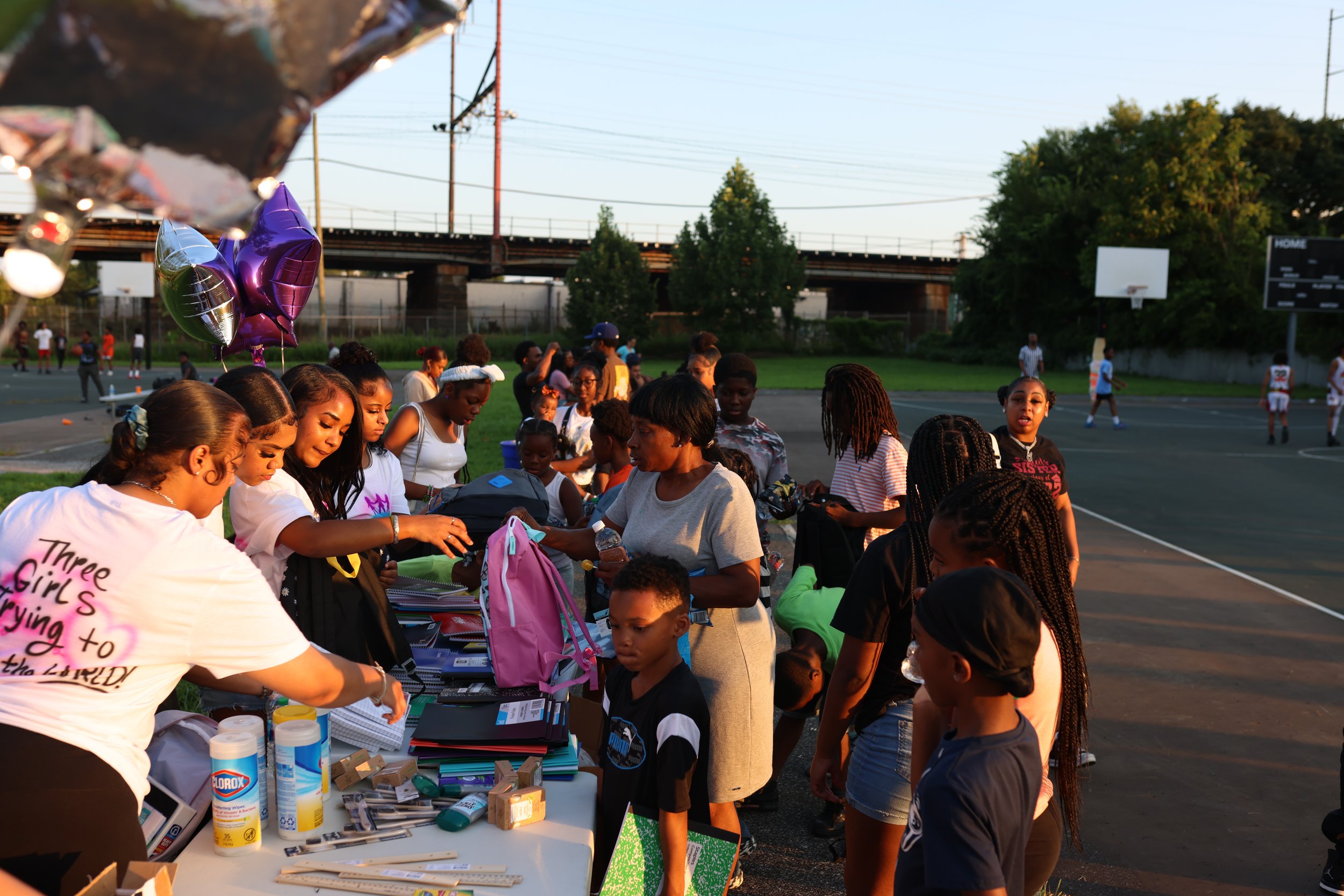 People line up at a table filled with school supplies and balloons at an outdoor event on a basketball court, with trees, a bridge, and a scoreboard visible in the background during late afternoon or early evening.