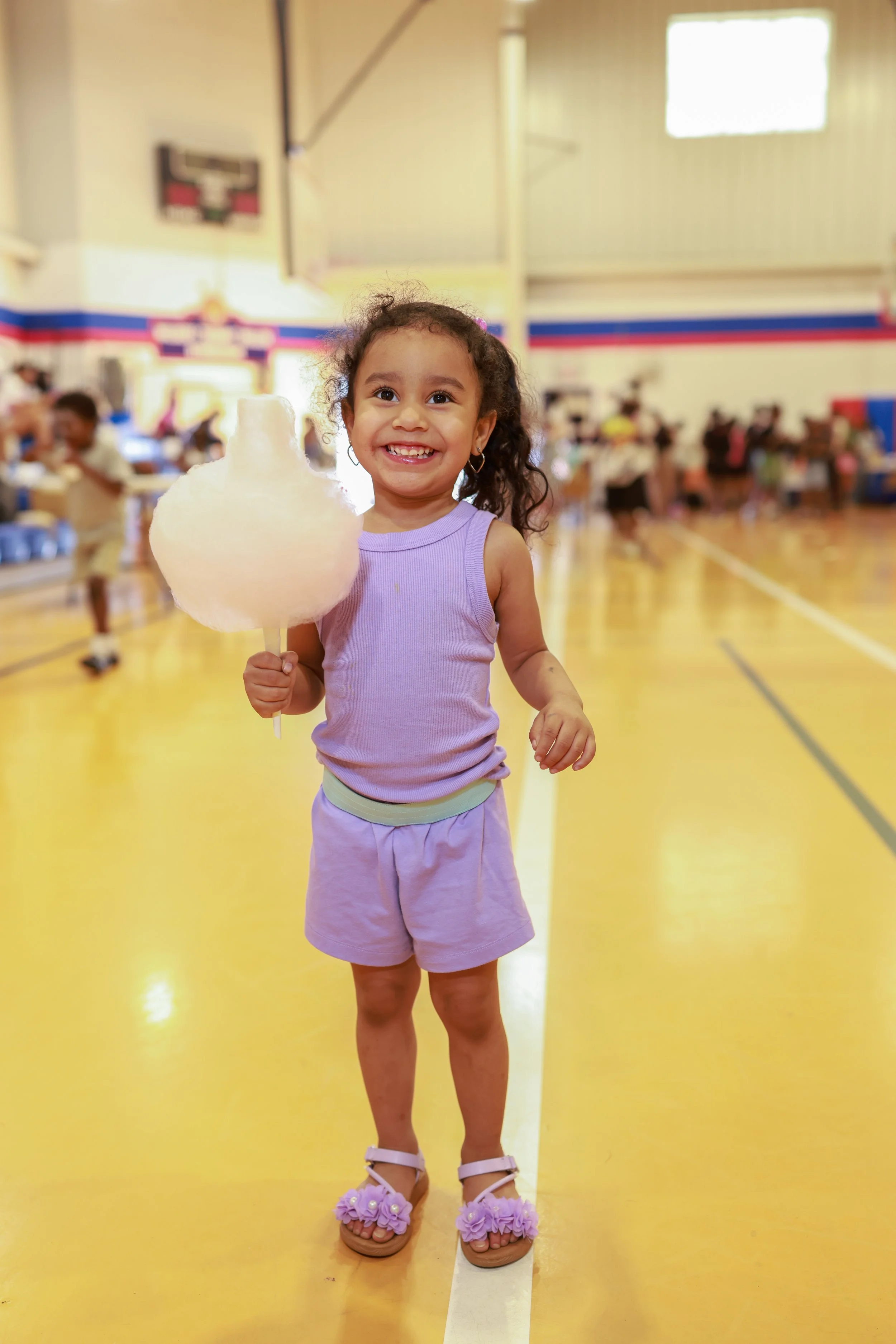 A young girl smiling, holding a large piece of cotton candy in an indoor gymnasium. She is wearing purple sports attire and sandals with purple ruffles. The background shows other children and adults in the gym.