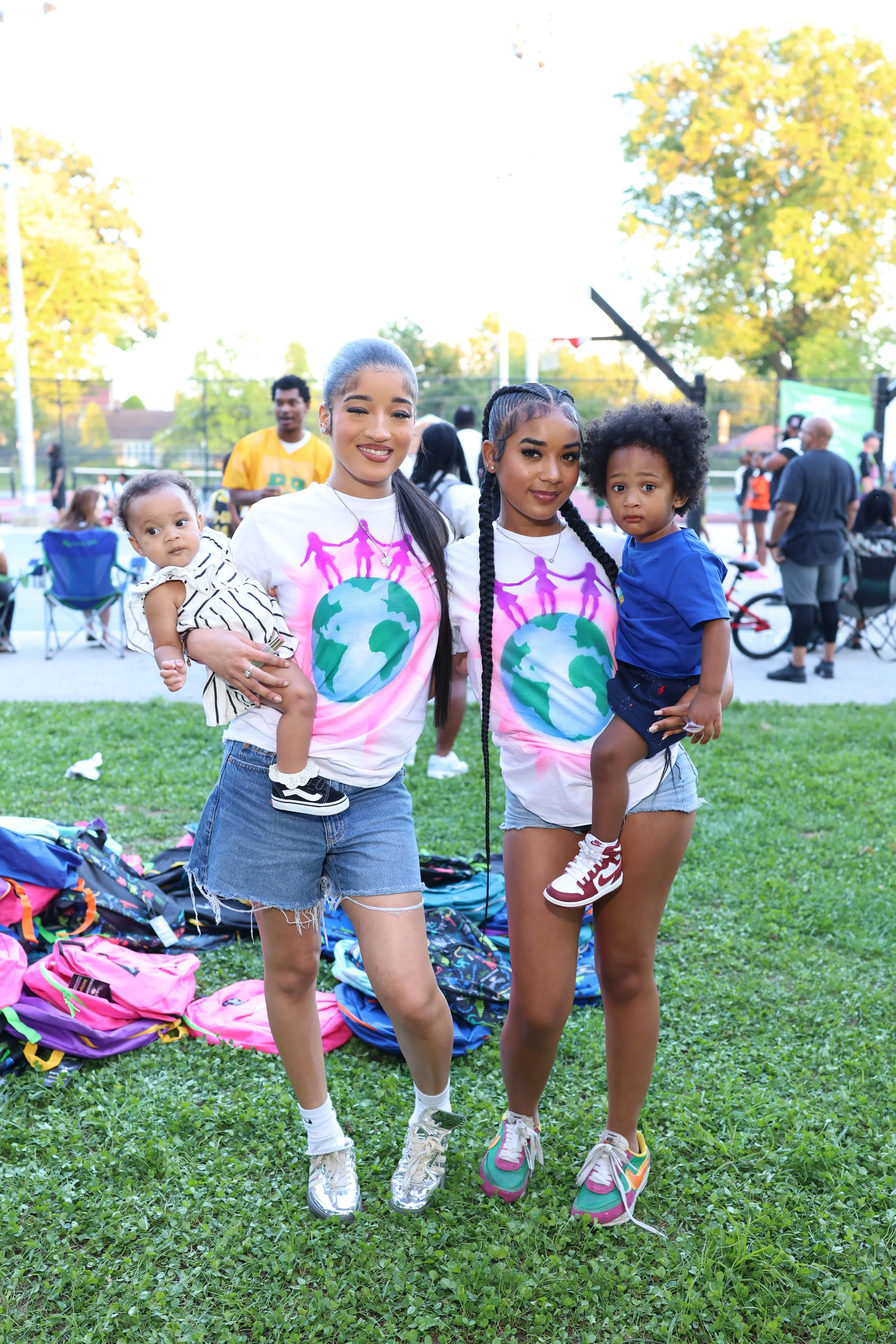 Two women holding children, standing on a grassy field at an outdoor event with scattered people, trees, and sports courts in the background.