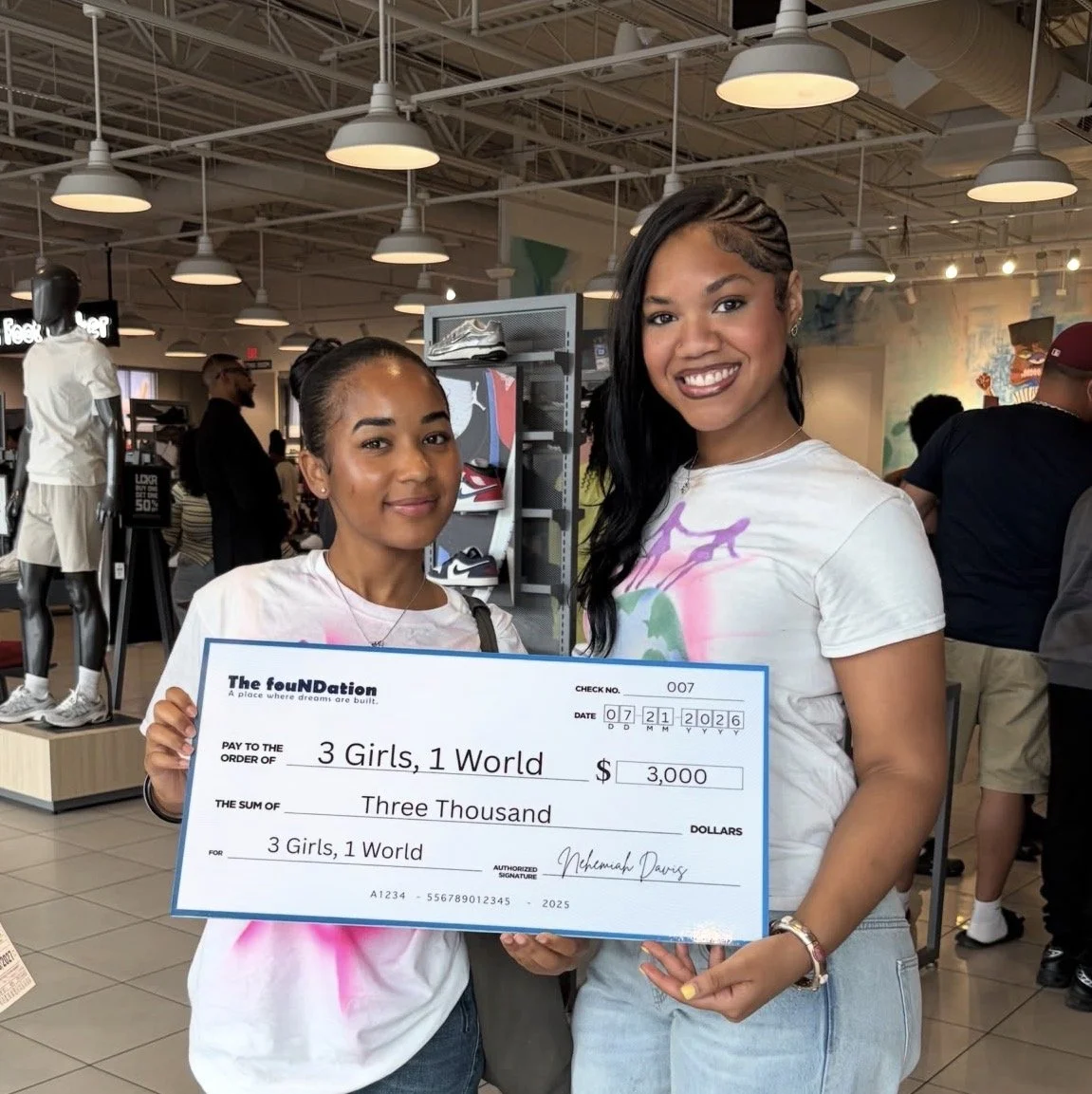 Two young women in a retail store, holding a large ceremonial check for $3,000 made out to 3 Girls, 1 World. Both women are smiling, with the woman on the left holding the check and the woman on the right standing beside her.