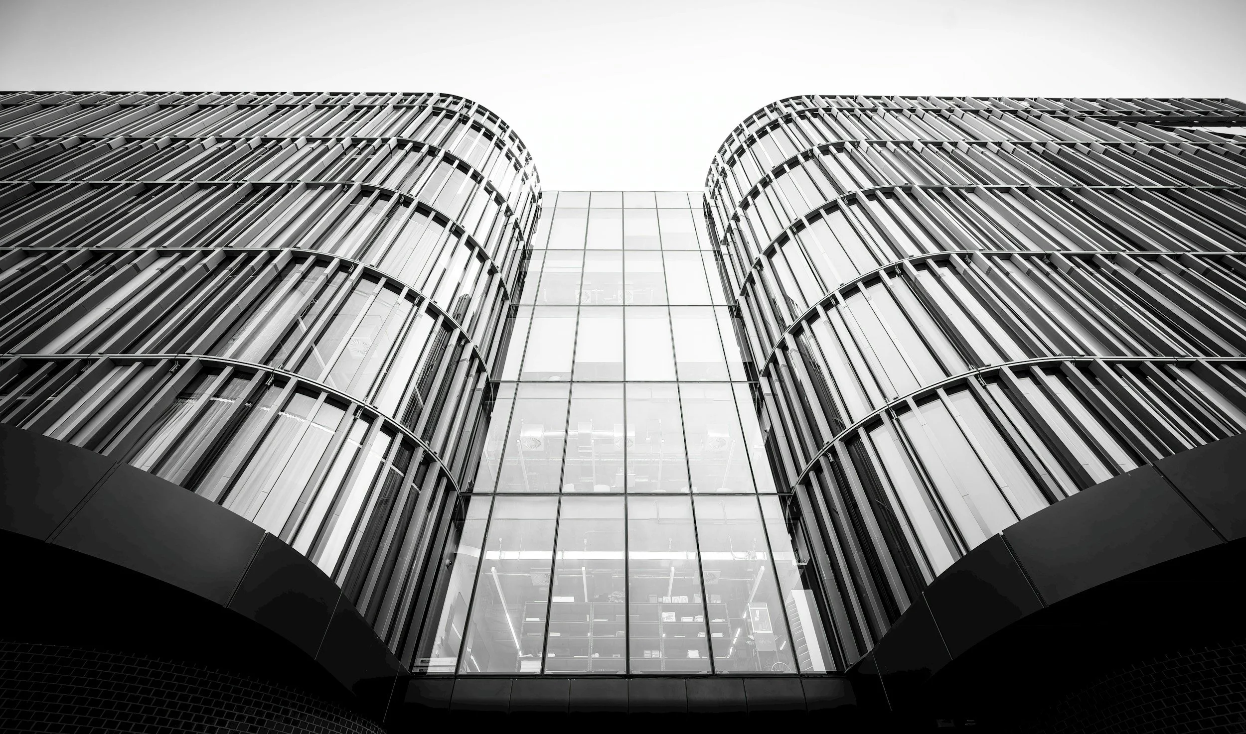 Low-angle black-and-white photograph of modern building with curved glass and metal facade, showing symmetrical architectural design.