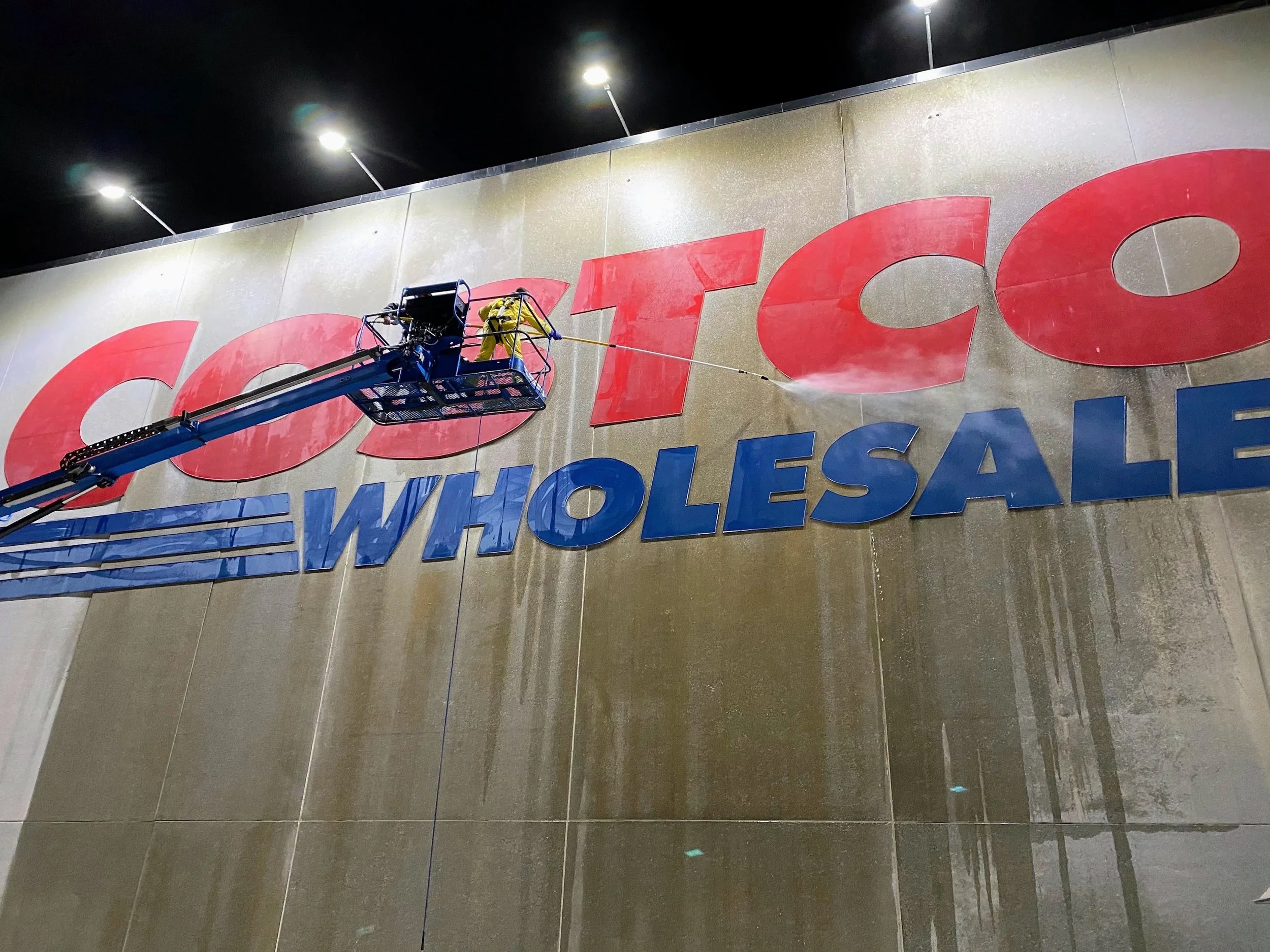 Worker on a lift cleaning a large sign on a building at night. The sign says 'GroceryWholesa' with some letters cut off.