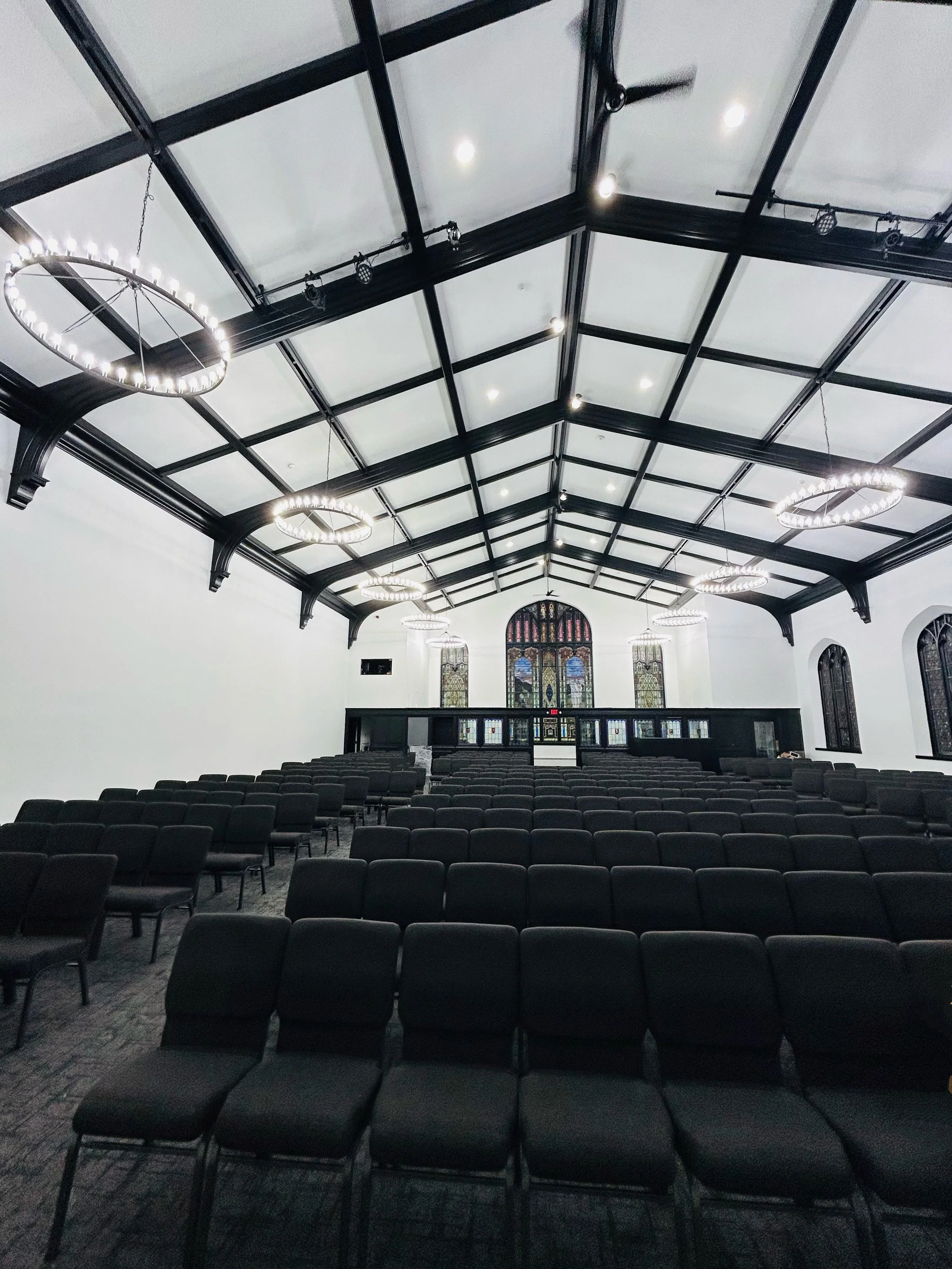 Empty auditorium with rows of black chairs, high ceiling with black beams, several large round chandeliers, stained glass windows, and a stage at the front.