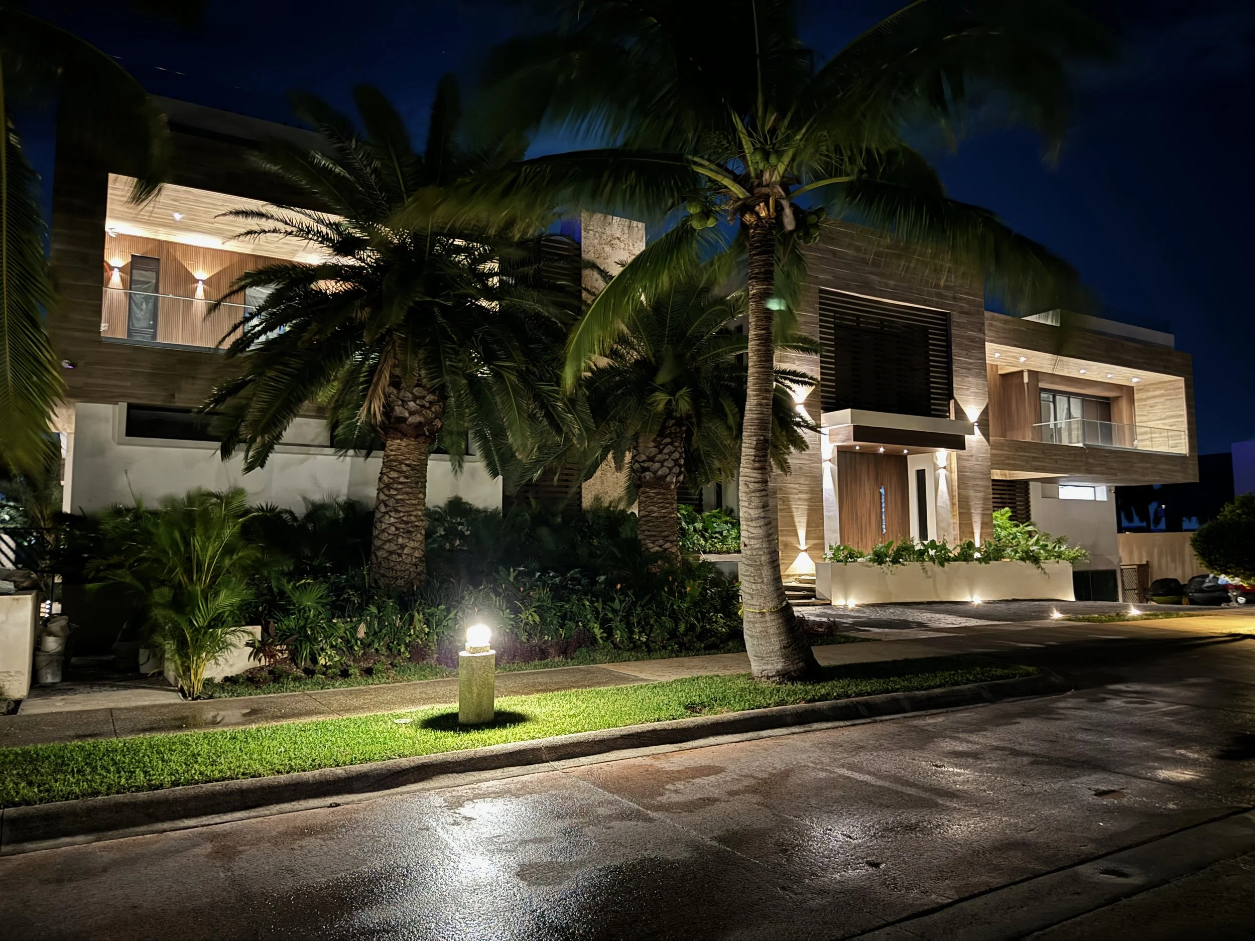 Night view of a modern residential building with exterior lighting, surrounded by palm trees, with a wet street in the foreground.