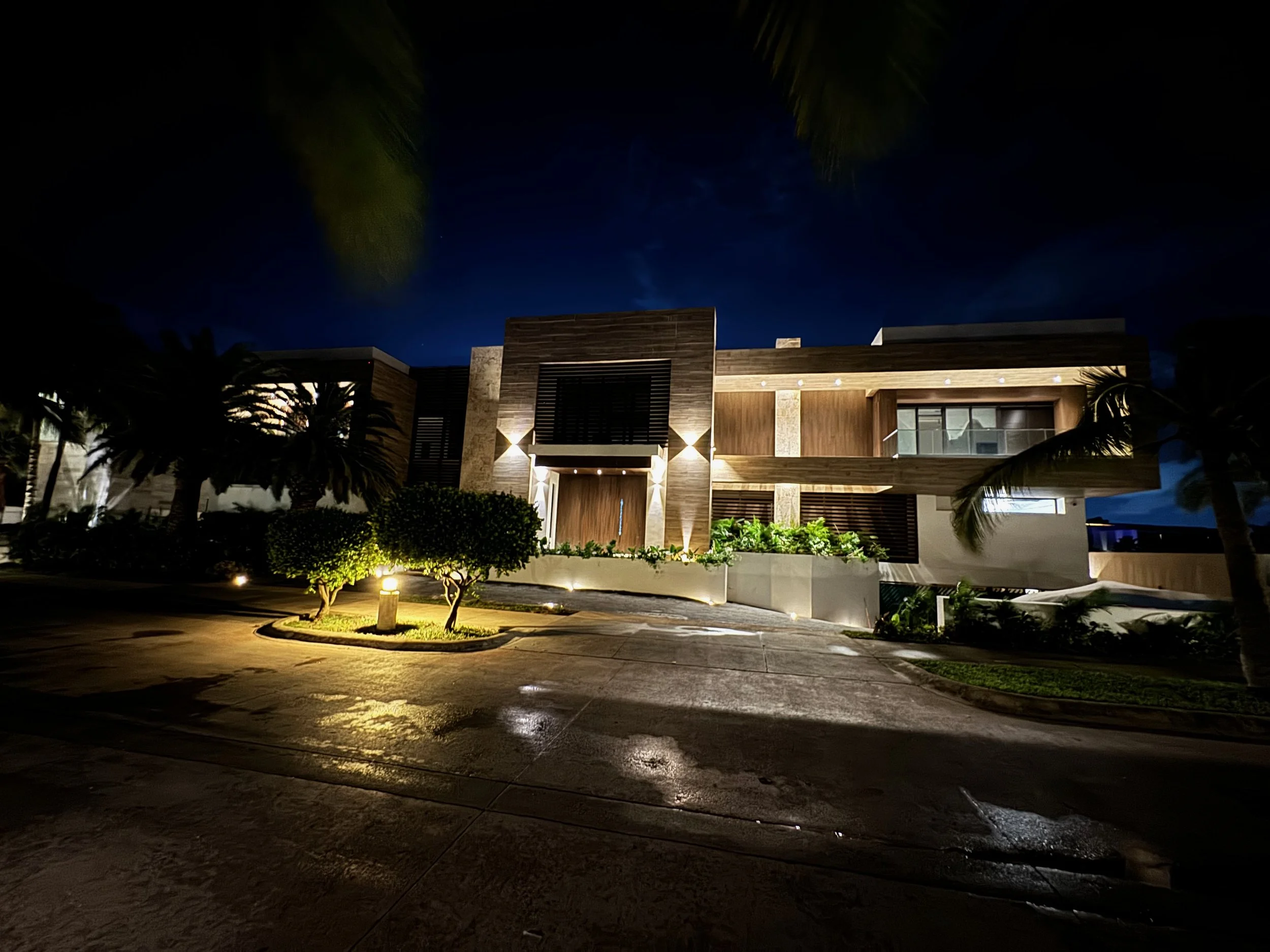 Nighttime view of a modern house with exterior lights, surrounded by palm trees and a driveway.