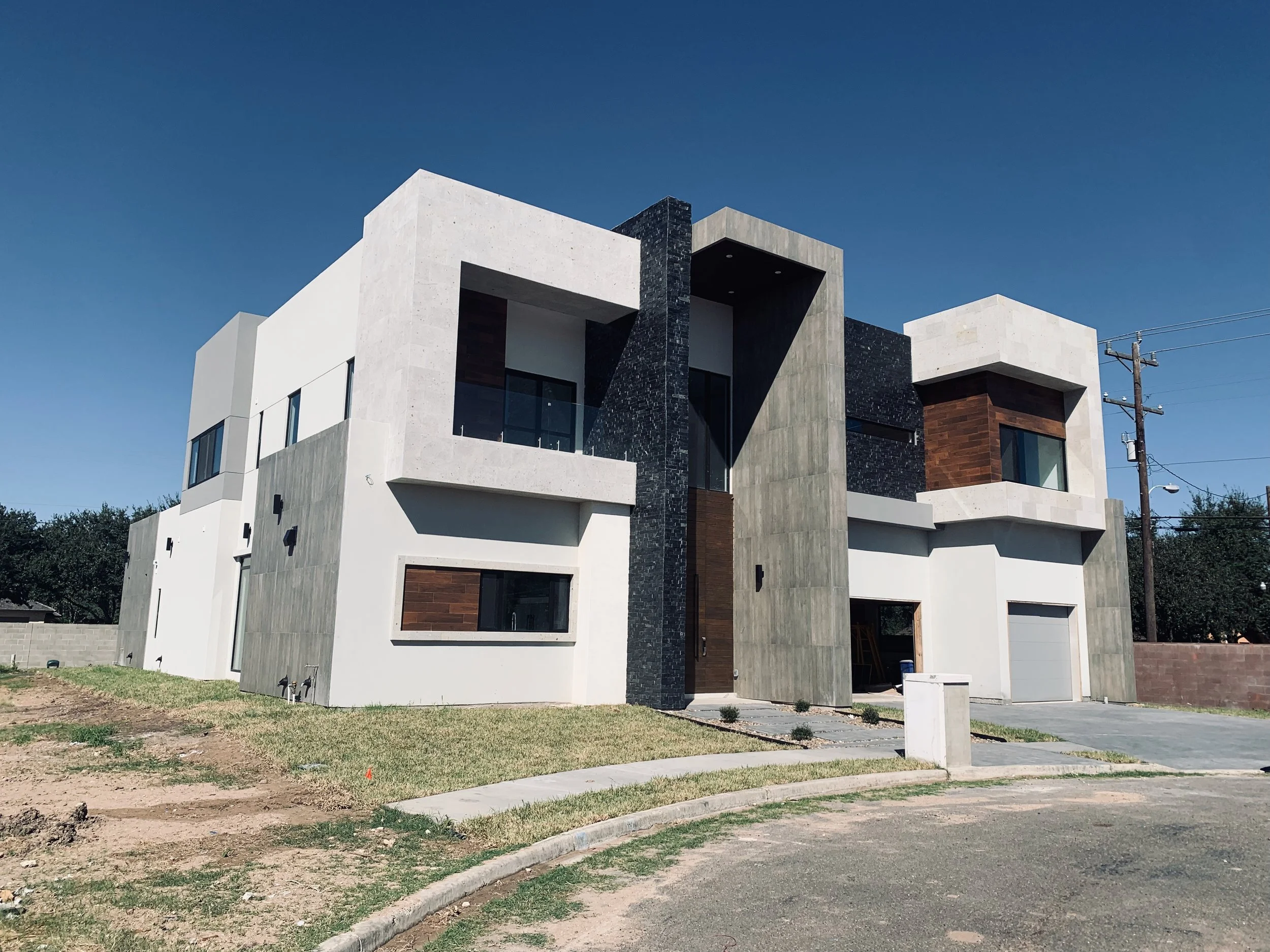 Modern multi-story house with white, black, and wooden exterior, large windows, and a small front lawn, with power lines in the background under a clear blue sky.