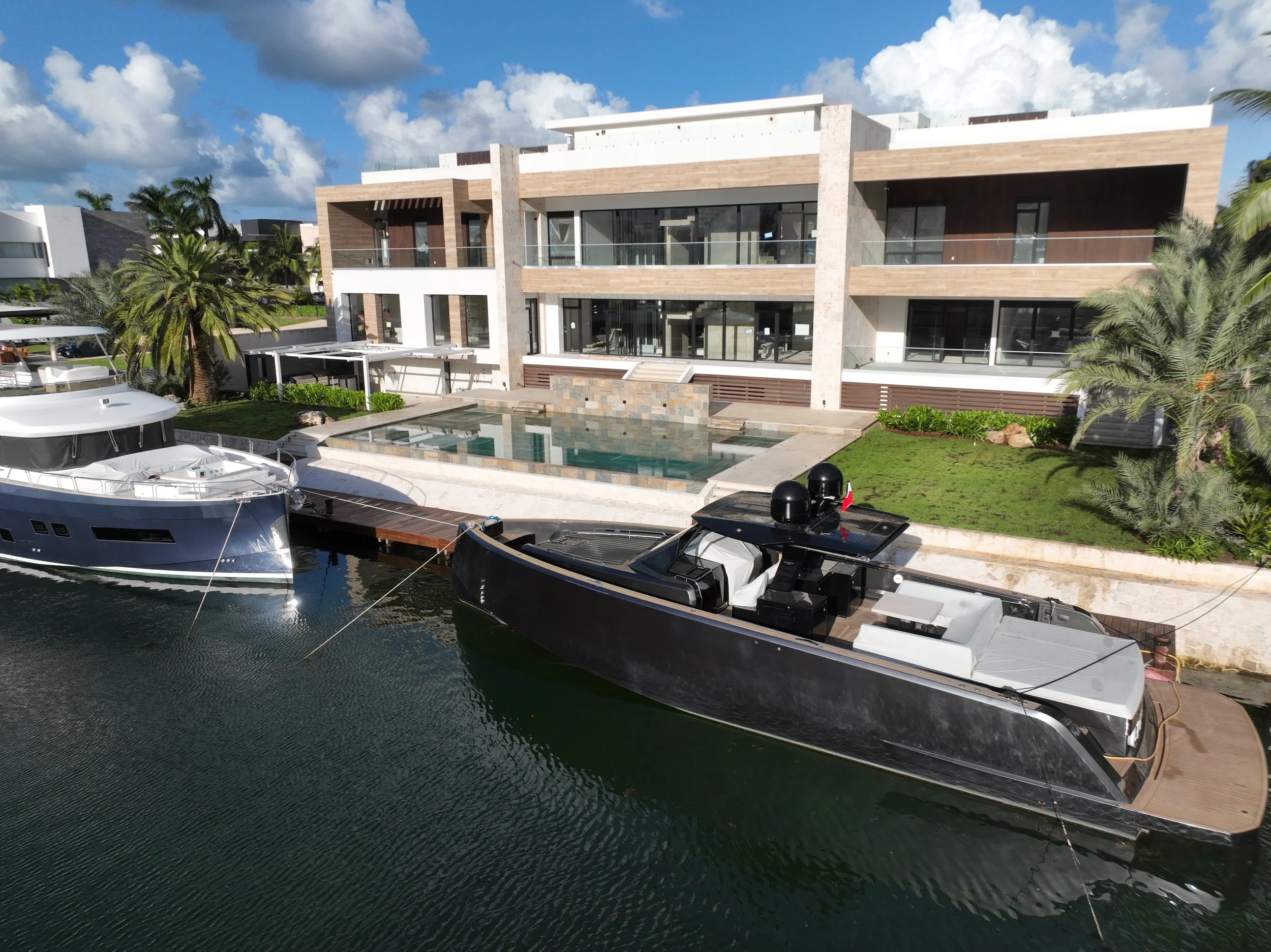Modern three-story waterfront house with large windows, balconies, and a swimming pool, surrounded by palm trees and moored boats in the water.