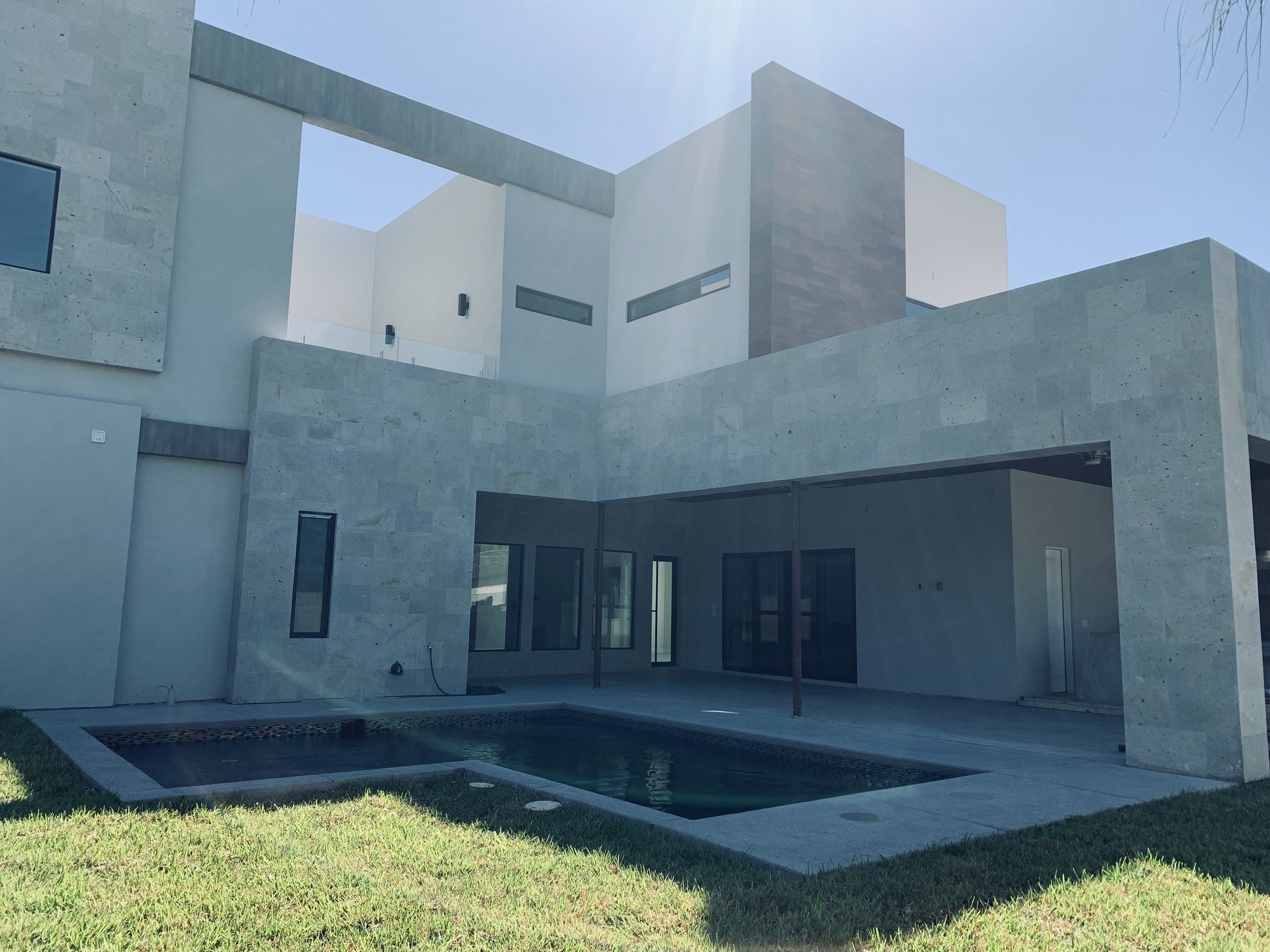 Modern house with clean lines and concrete exterior, large windows, a patio area, a small pool, and a green lawn under a clear sky.