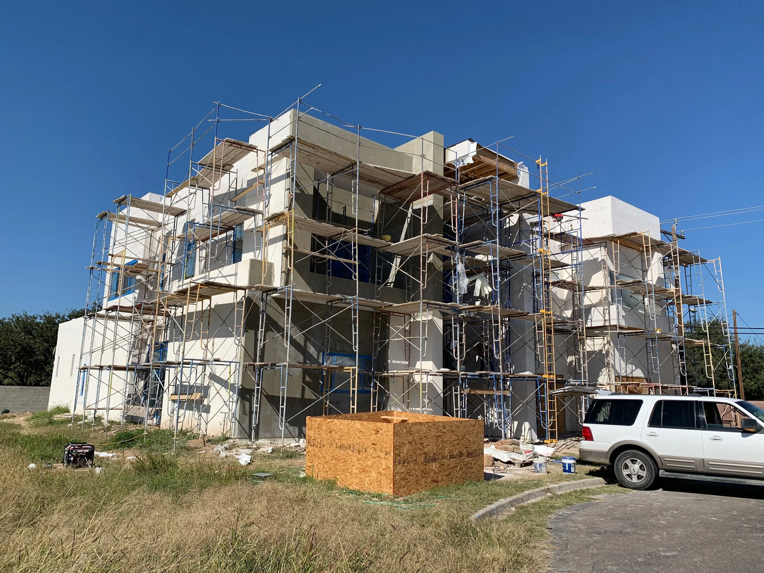 Construction site of a multi-story building with scaffolding, building materials, and a parked white vehicle in the foreground, under a clear blue sky.