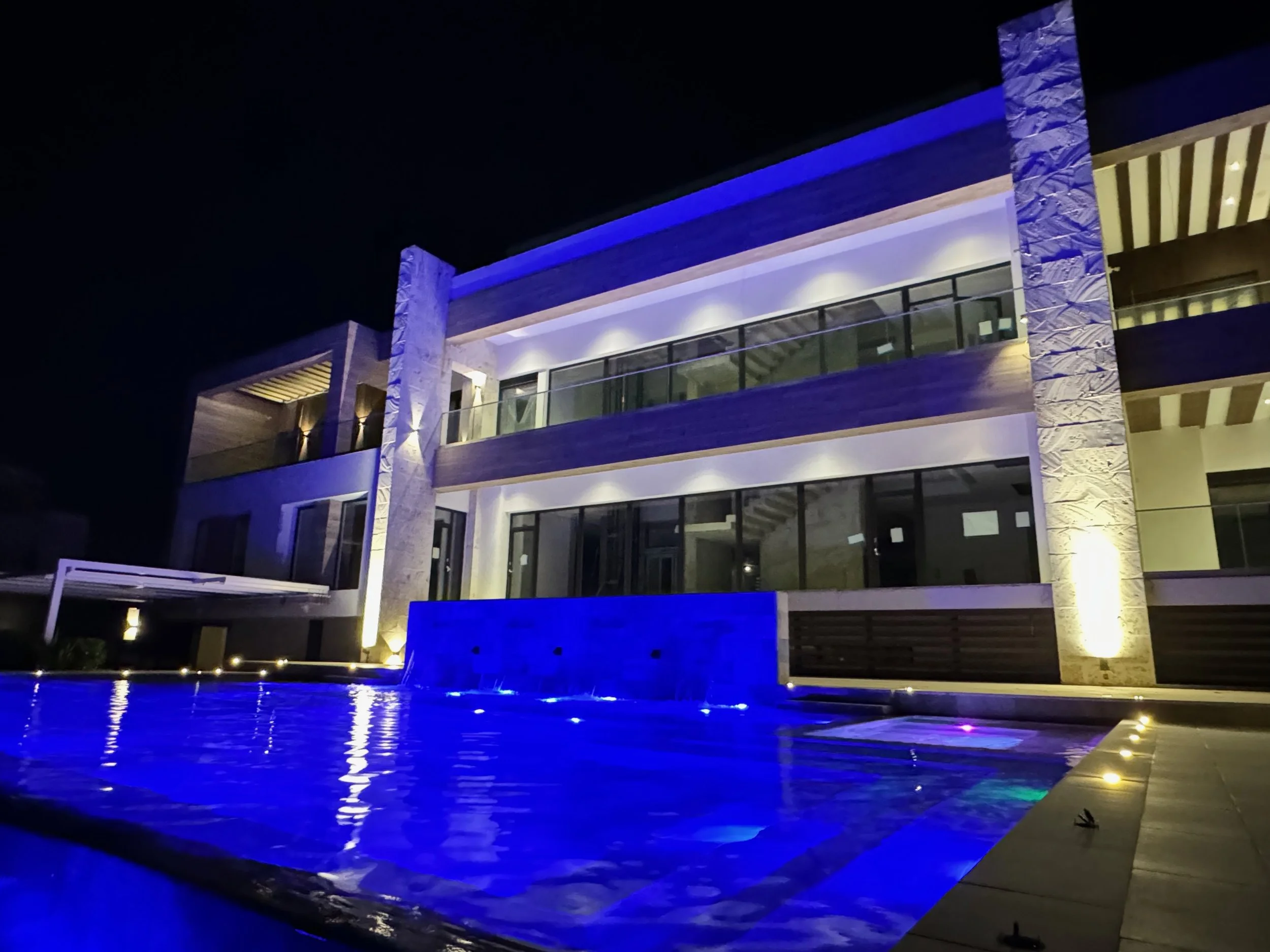 Modern multi-story building at night with illuminated balconies and blue accent lighting, adjacent to a pool reflecting the building's lights.