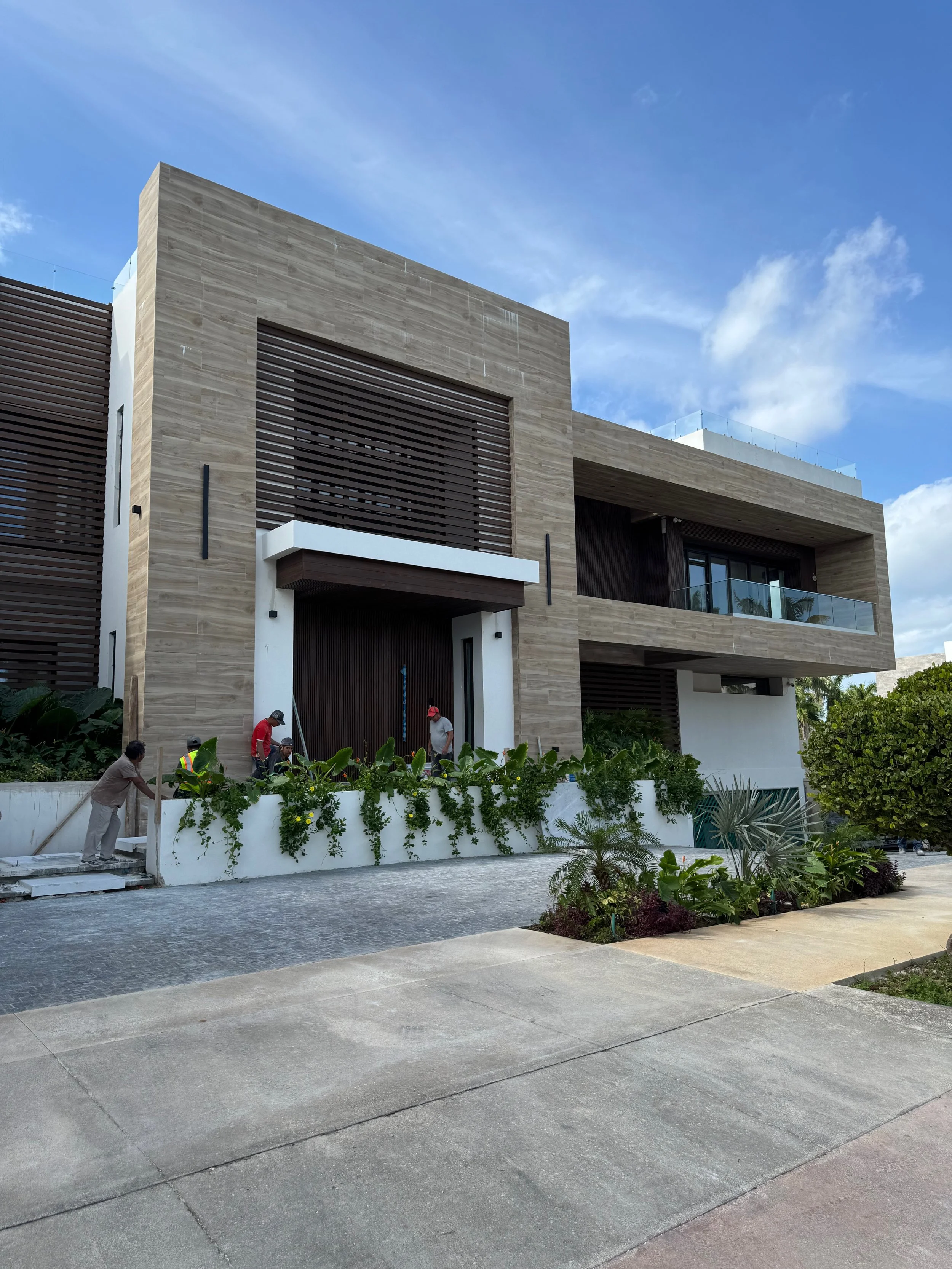 A modern multi-story house with a neutral-colored facade, balcony with glass railings, and horizontal window shutters. There are workers in the front yard planting and maintaining plants, and palm trees and other greenery are visible around the area. The sky is blue with some clouds.