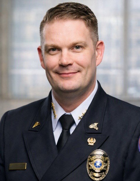 A smiling man in a police uniform with badge and insignia, standing in front of a window with blurred cityscape background.