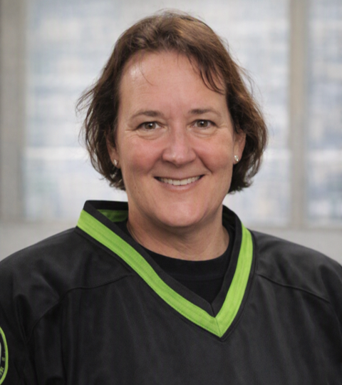 A woman with short brown hair smiling, wearing a black sports jersey with green trim, standing in front of a window with blinds.