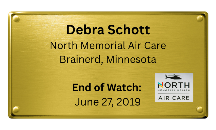 Gold memorial plaque with black text, dedicated to Debra Schott of North Memorial Air Care in Brainerd, Minnesota, marking her end of watch on June 27, 2019, featuring the North Memorial Health Air Care logo with a helicopter icon.