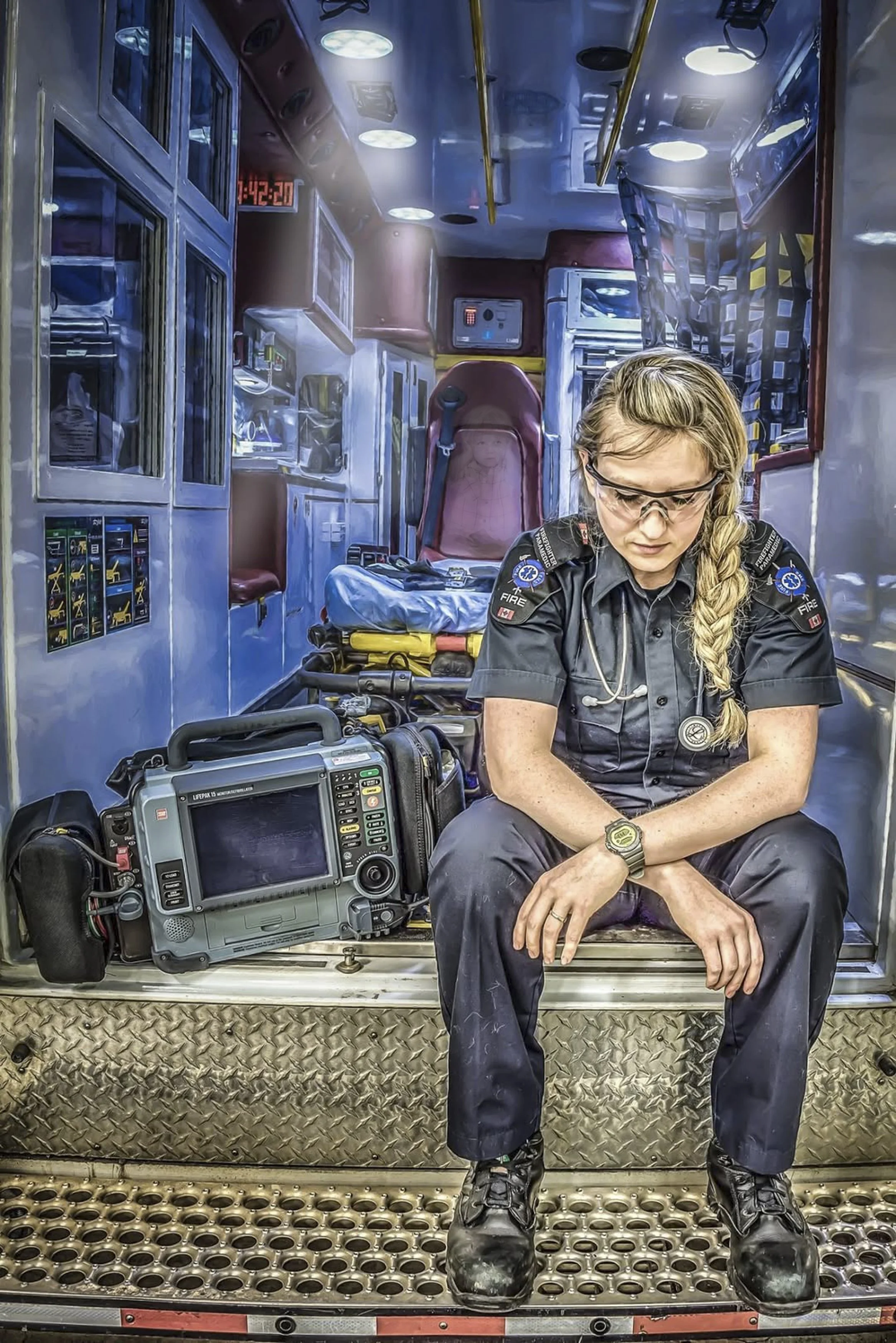 A female paramedic sitting inside an ambulance with medical equipment, a defibrillator, and a monitor around her, looking down with sunglasses and braided hair.