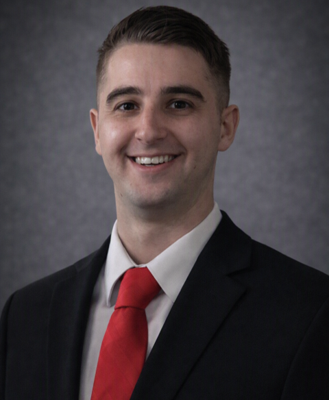Portrait of a young man in a black suit, white shirt, and red tie, smiling against a gray background.