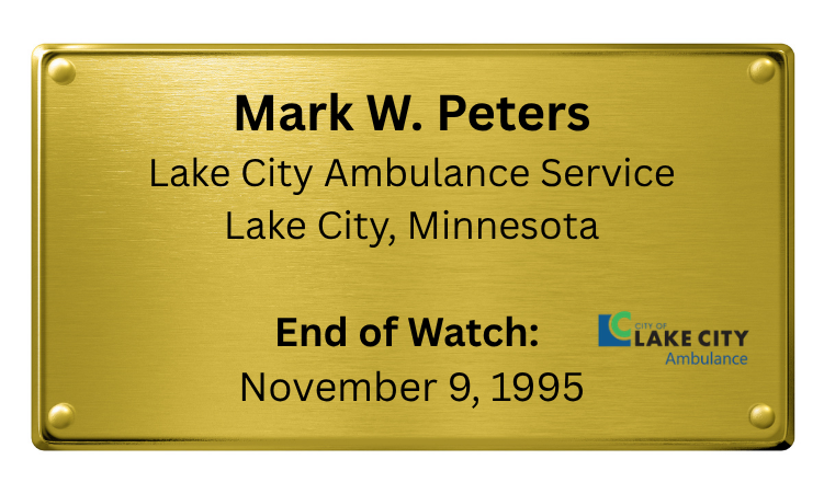 A gold-colored metal plaque with black text and a small logo in the lower right corner. The plaque reads: "Mark W. Peters, Lake City Ambulance Service, Lake City, Minnesota. End of Watch: November 9, 1995."