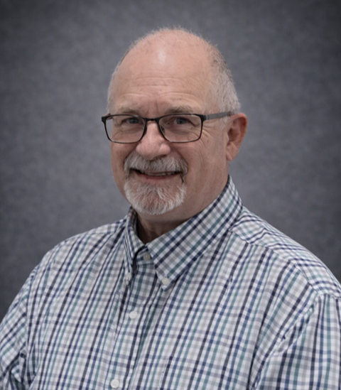 A middle-aged man with glasses and a beard, wearing a plaid button-up shirt, smiling against a gray background.