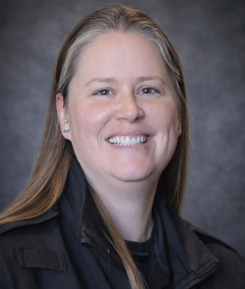 Portrait of a smiling woman with long blonde hair, wearing a black jacket and earrings, against a gray background.
