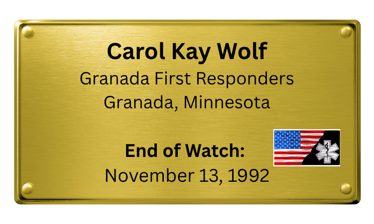 A gold-colored plaque with black text honoring Carol Kay Wolf, a first responder from Granada, Minnesota, indicating the end of a watch on November 13, 1992, with an American flag and medical symbol in the lower right corner.