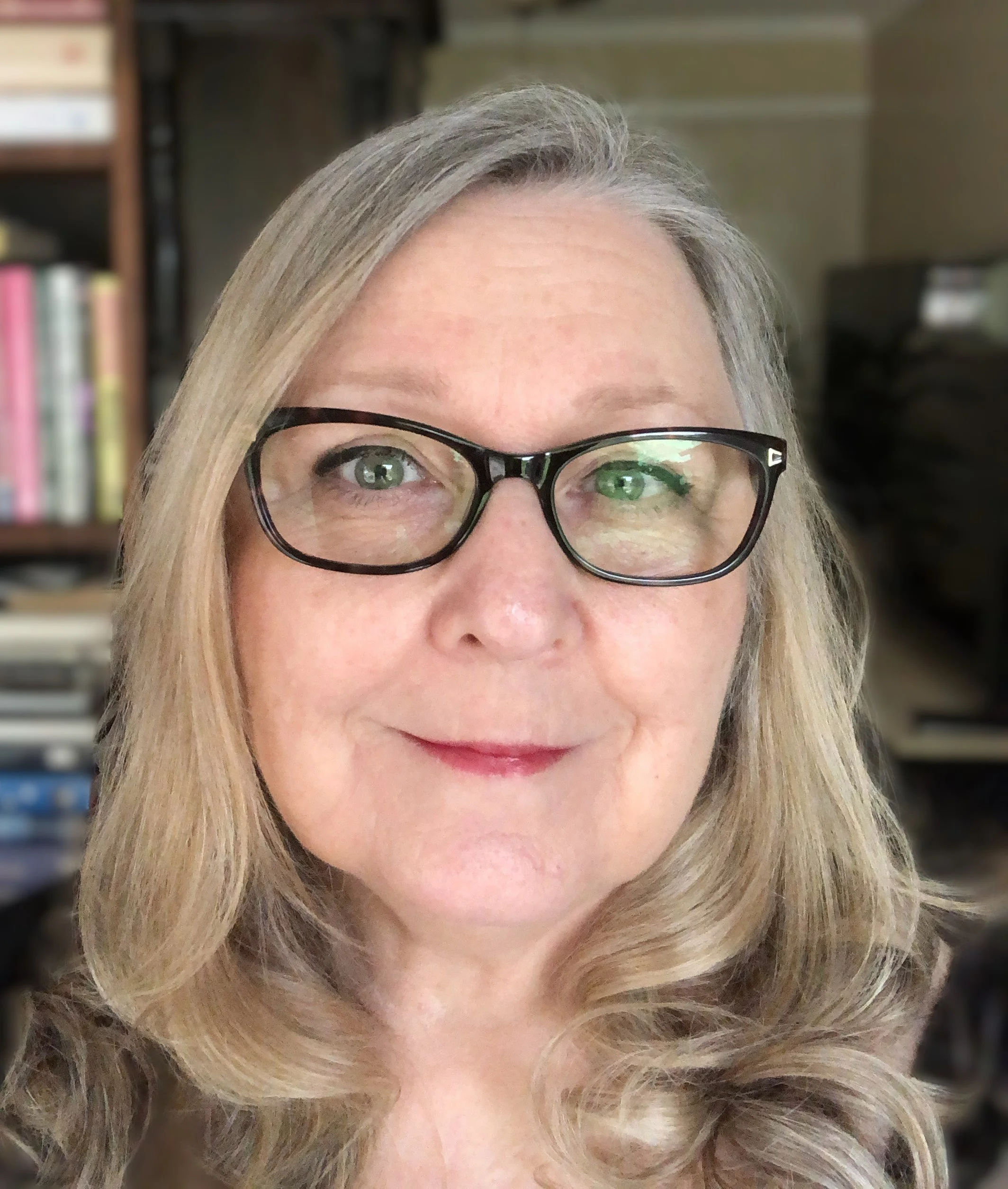 Close-up of a woman, the artist Theresa Parsons, with blonde hair and black glasses, showing her face and hair against a blurred background with shelves.