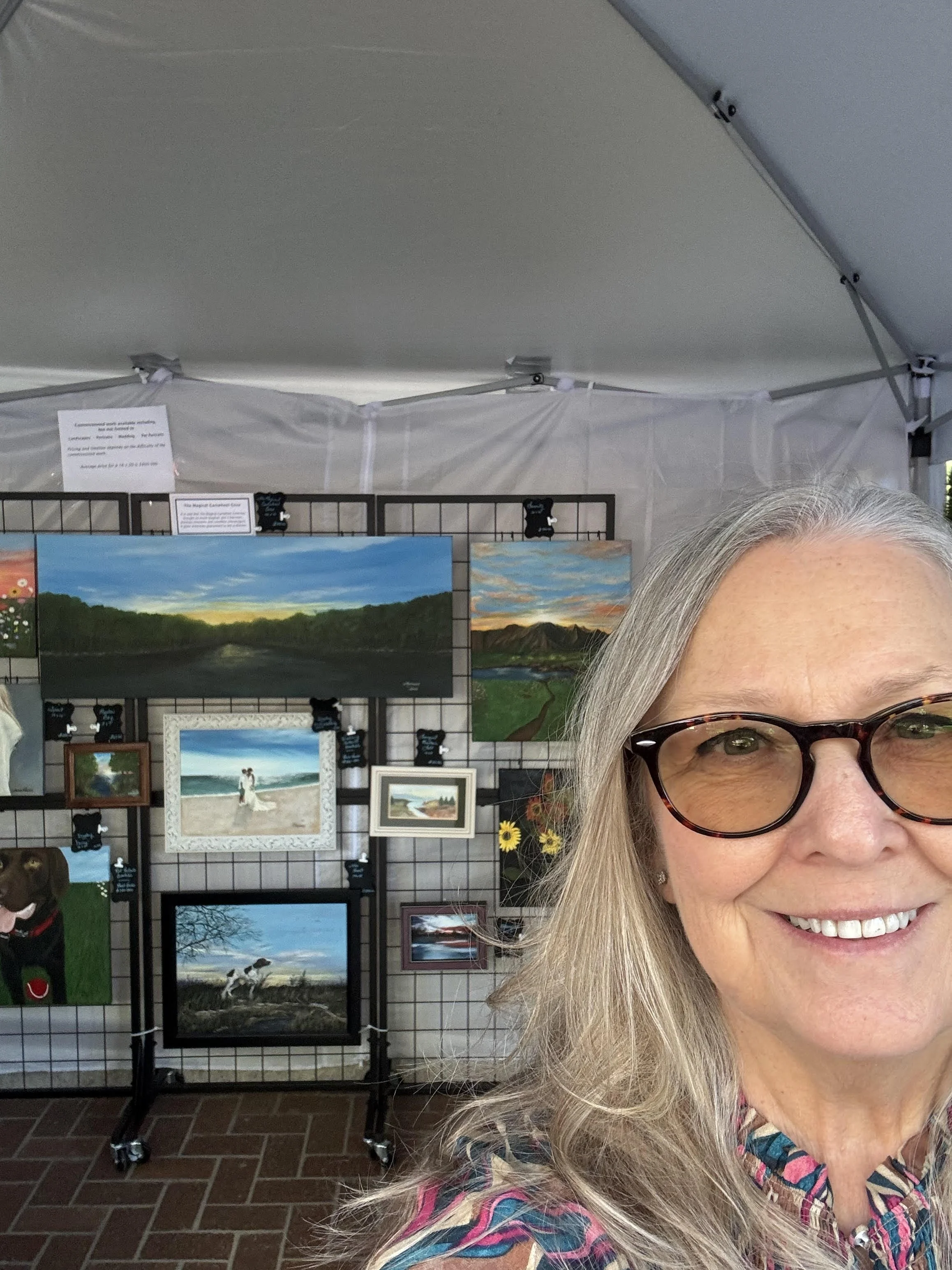 Partially visible image of the artist, Theresa Parsons, a woman with gray hair, glasses, a colorful patterned shirt, smiling, in front of a display of landscape paintings at an art exhibit.