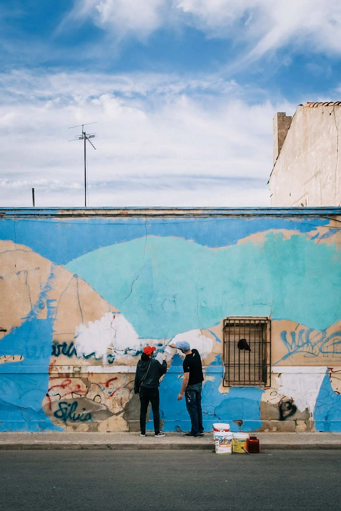 Dos personas pintando un mural en una pared de colores claros, una con gorra roja y la otra con gorra azul, frente a un cielo con nubes.