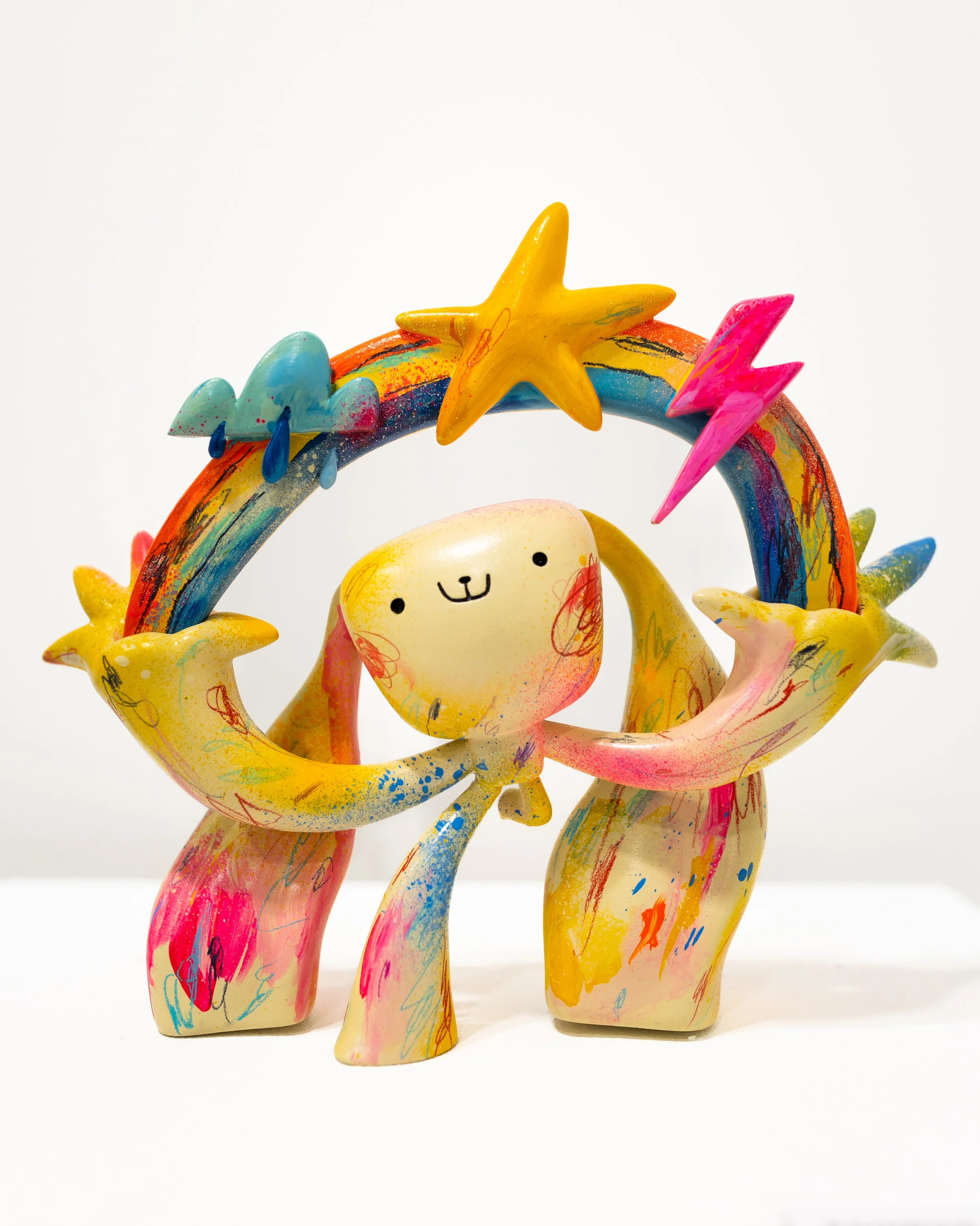 Escultura colorida de figuras tipo art toy, estrellas, arco iris y gotas, con una cara de cartoon sonriente en el centro.