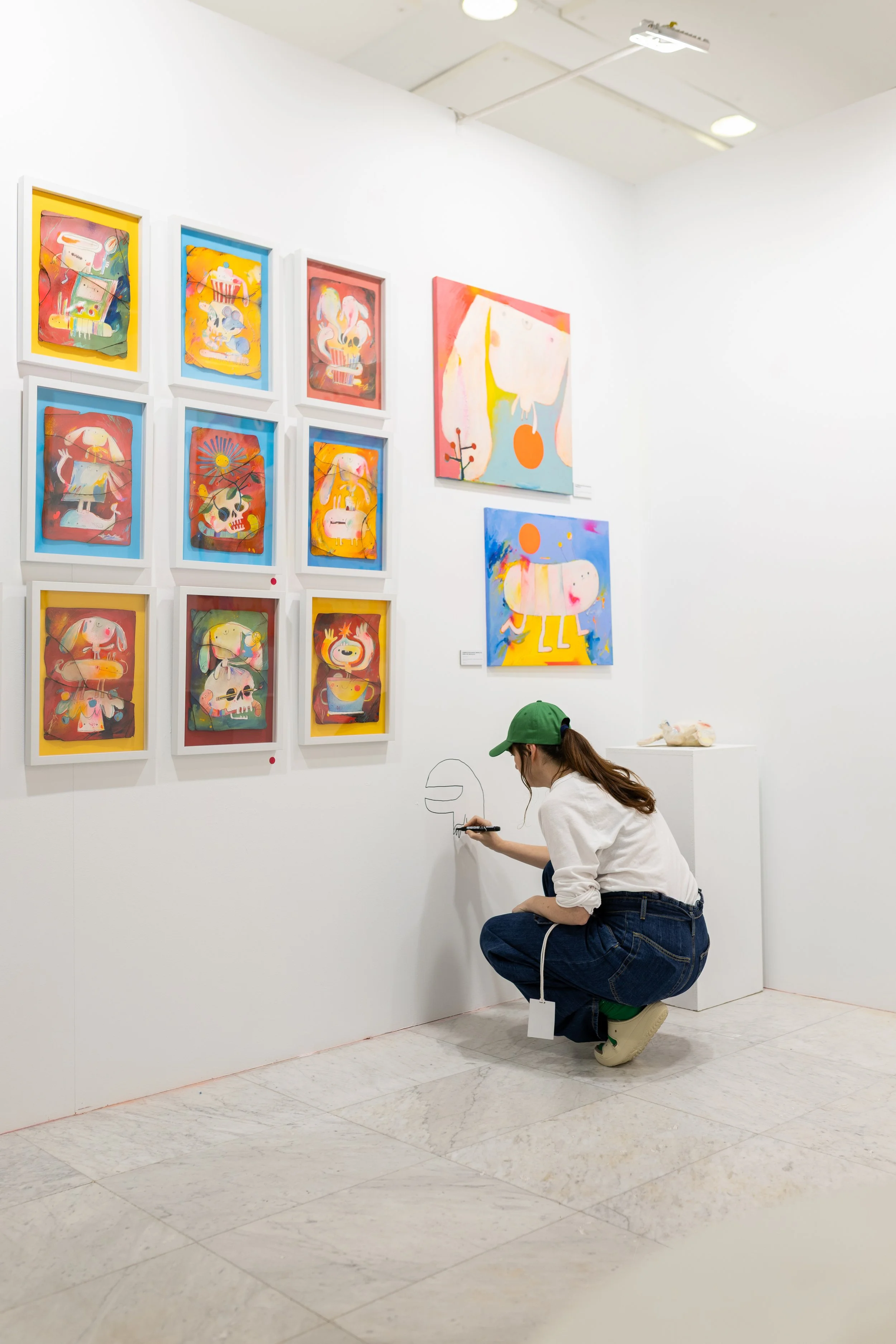 Una persona joven, con gorra verde, camiseta blanca y pantalones vaqueros, está agachada pintando en la pared de una galería de arte. En la pared hay varias obras de arte coloridas y abstractas, algunas enmarcadas y otras no.