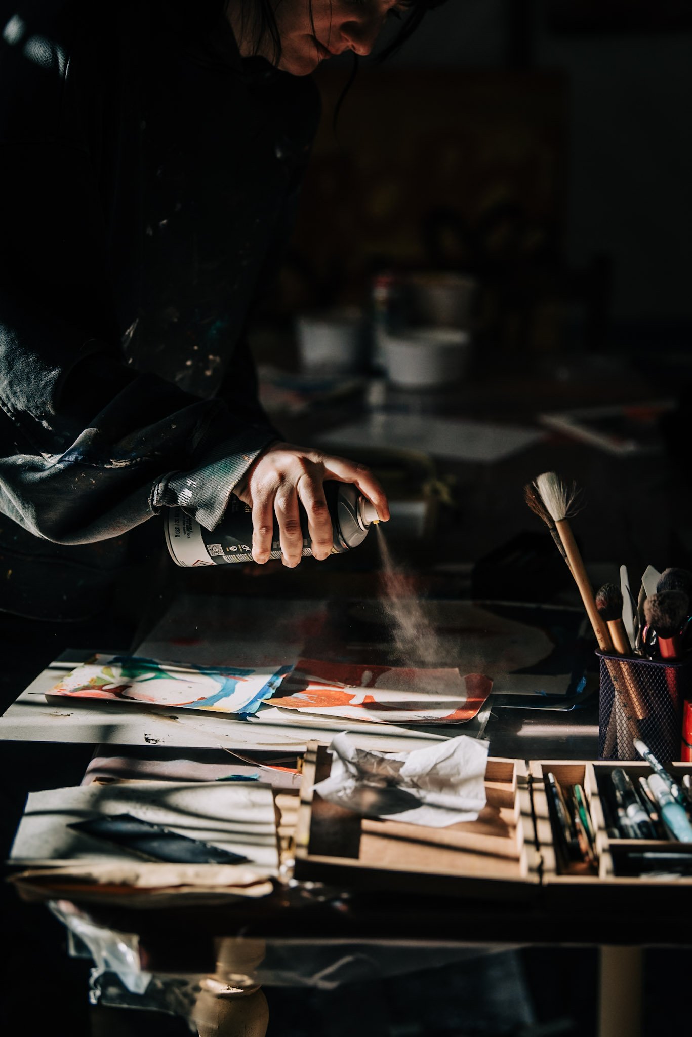 Persona pintando con aerosol en un espacio oscuro, rodeada de papeles y utensilios de arte en una mesa