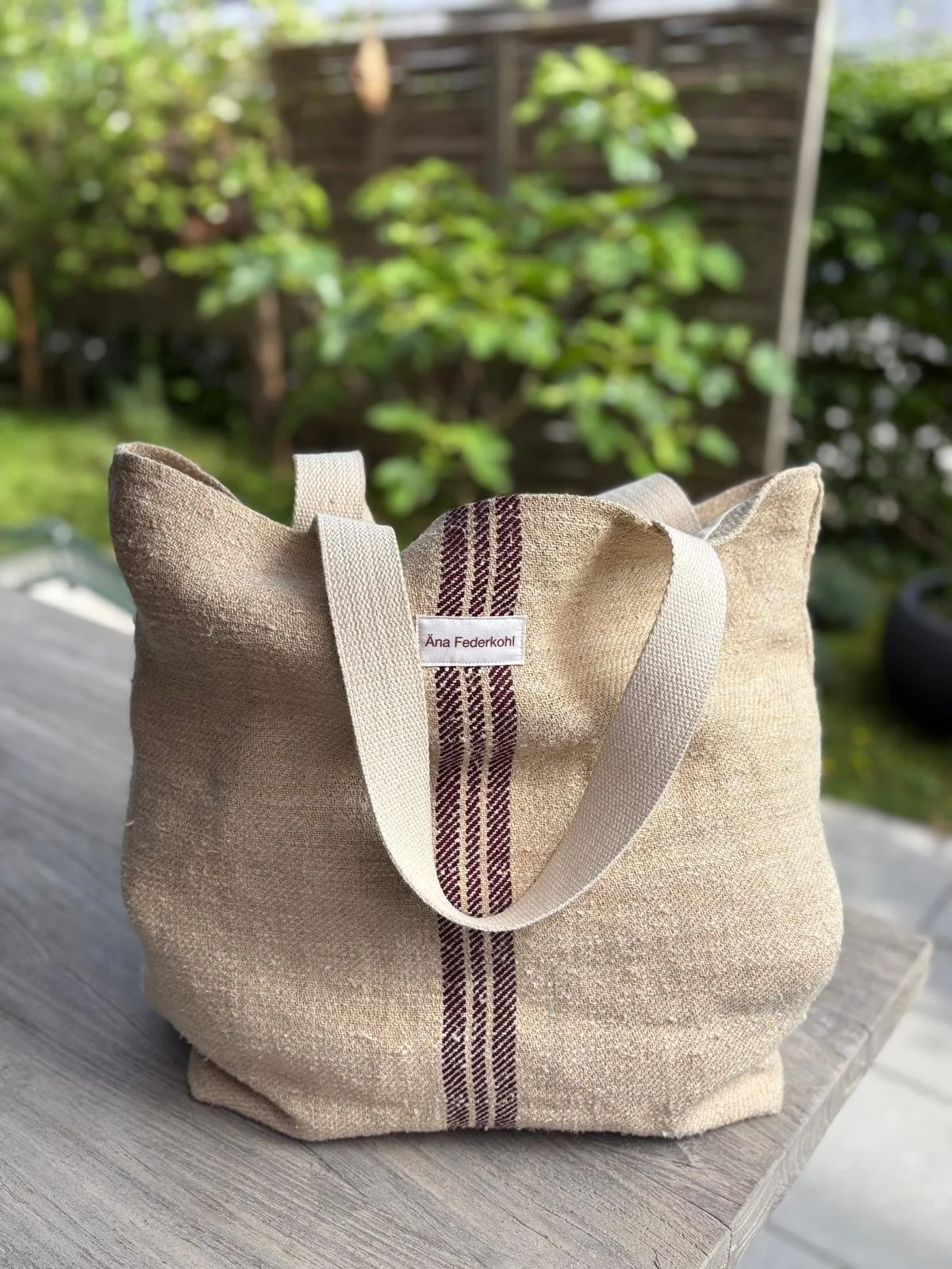 Eine beige Stofftasche mit roten Streifen und dem Label 'Ána Federkohl' auf einer Holzplatte, im Hintergrund ein grüner Garten.