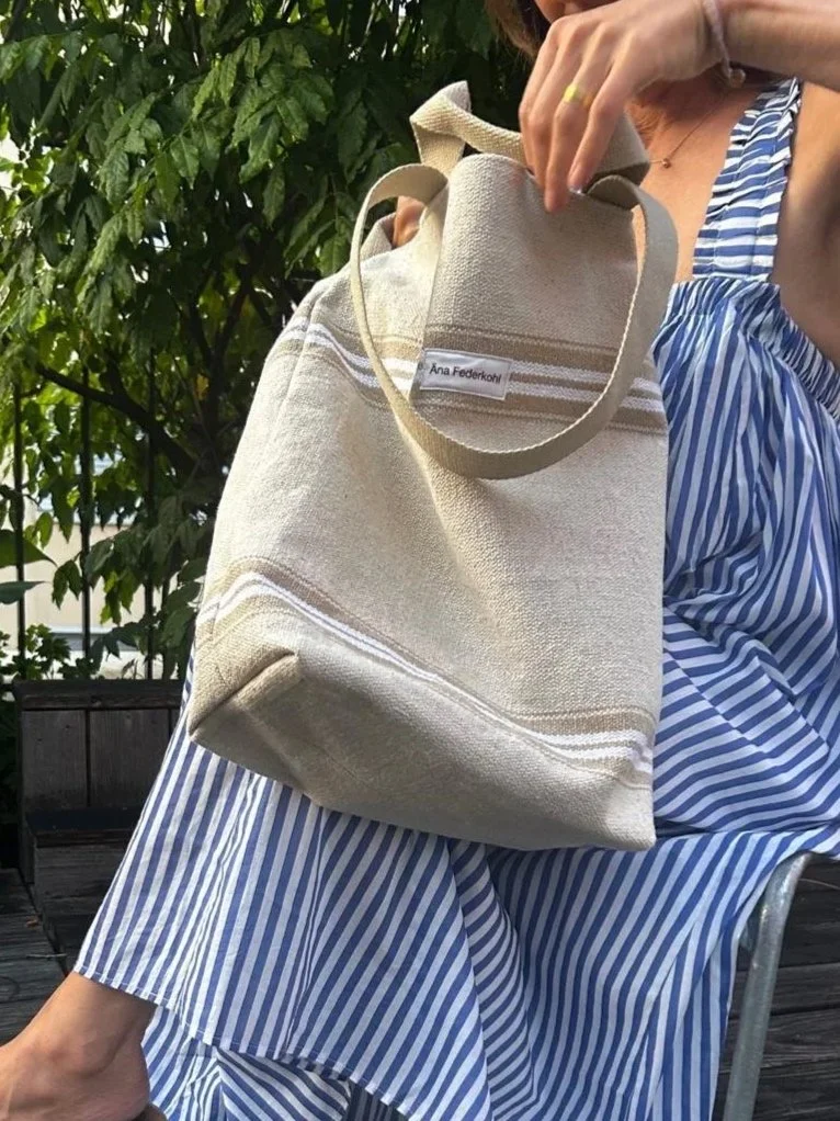 Eine Person hält eine große beige Stofftasche mit handgeschriebener Etikette, die auf 'Ana Federkoh' lautet. Die Person trägt einen blauen und weißen gestreiften Kleidungsstil. Im Hintergrund sind grüne Blätter sichtbar.