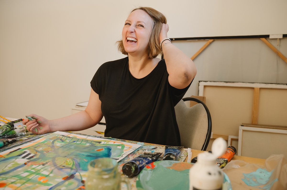 A woman in a black shirt laughing while sitting at a table covered with art supplies and colorful paintings.